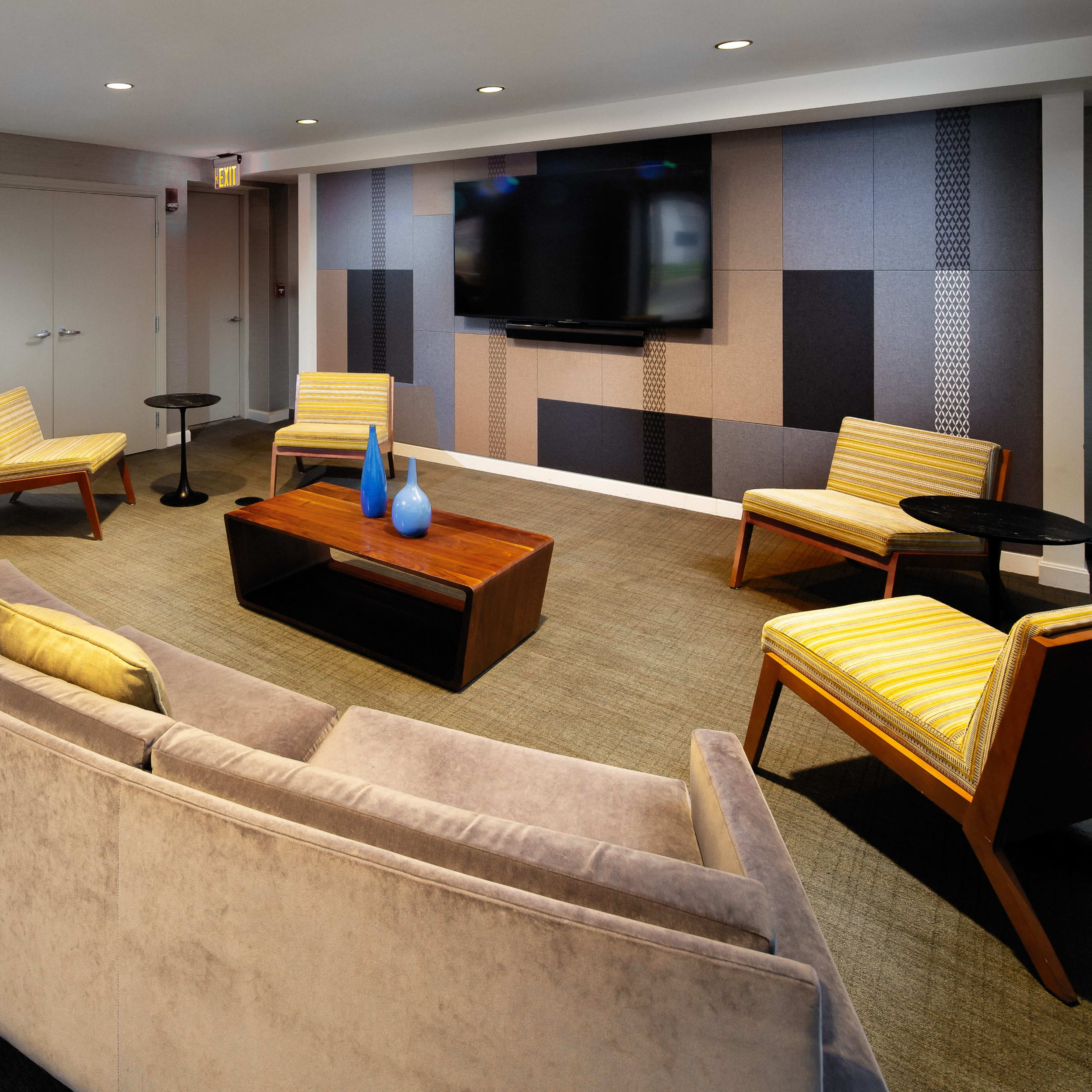 A modern lounge area featuring a curved gray sofa, several yellow-striped chairs, a wooden coffee table, and a wall-mounted television surrounded by patterned panels.