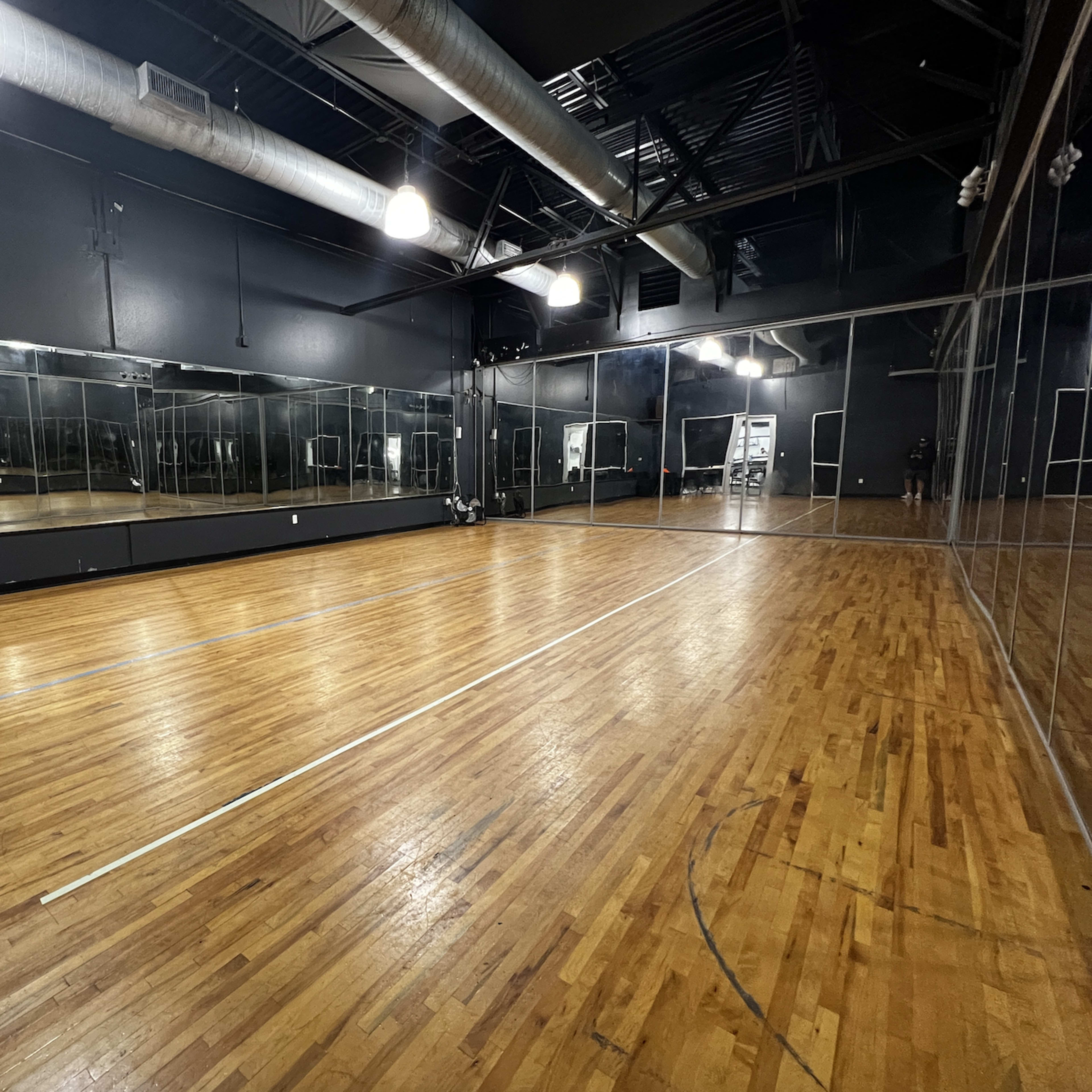 A spacious dance studio with wood flooring, large mirrors along one wall, and overhead lighting.