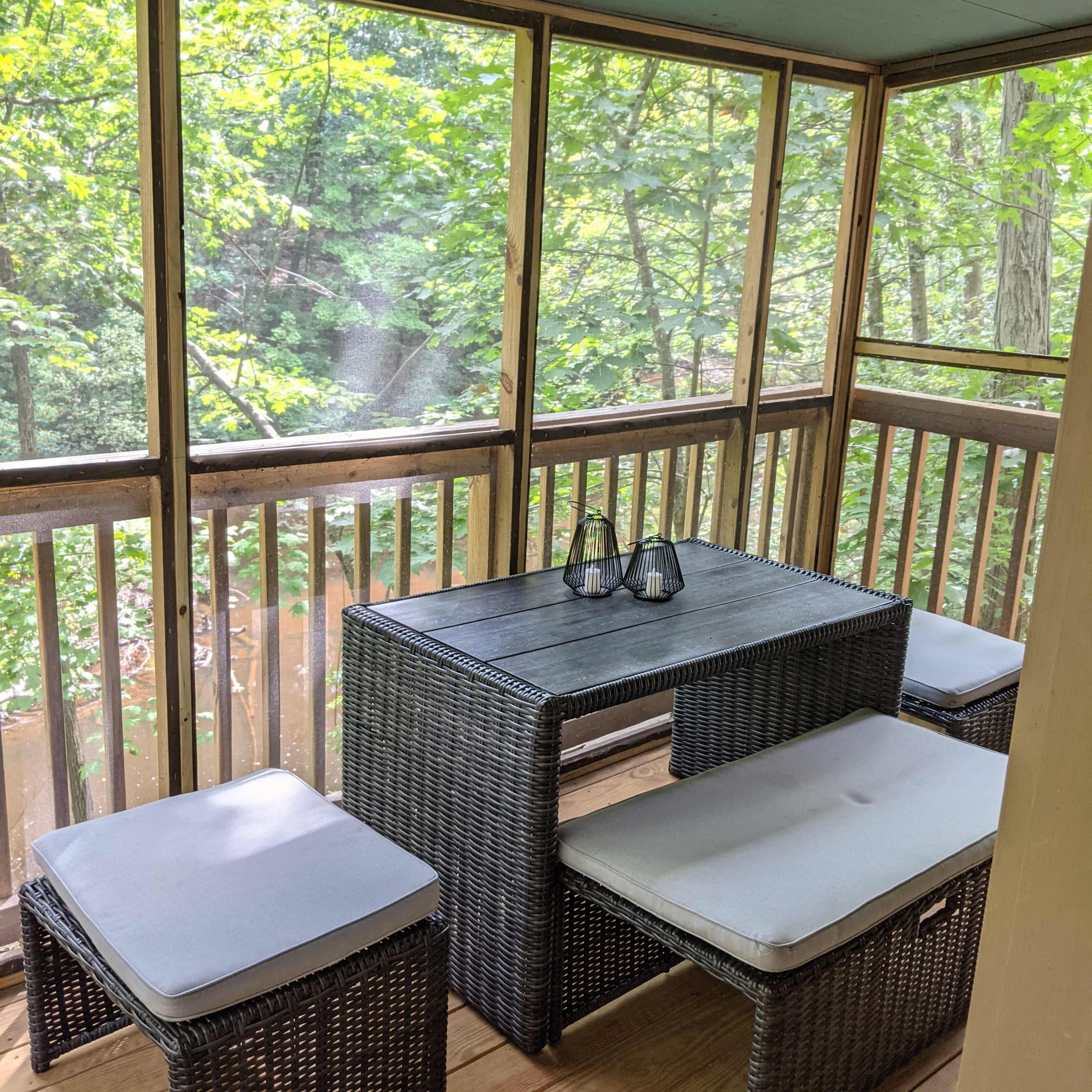 A screened porch features a rectangular table surrounded by four cushioned benches, with greenery visible through the screens.