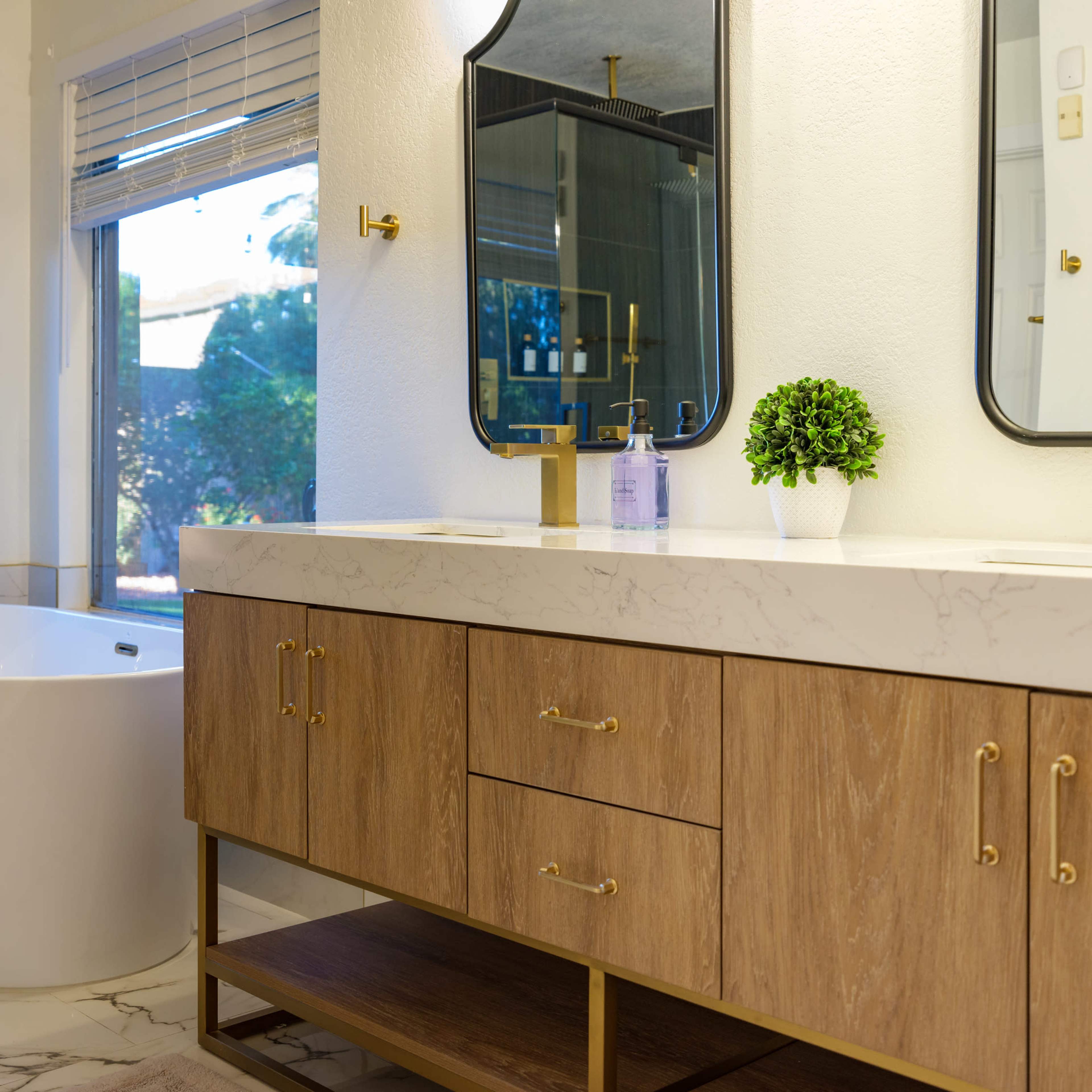 A modern bathroom with a freestanding bathtub, a double vanity with gold fixtures, and large windows allowing natural light.