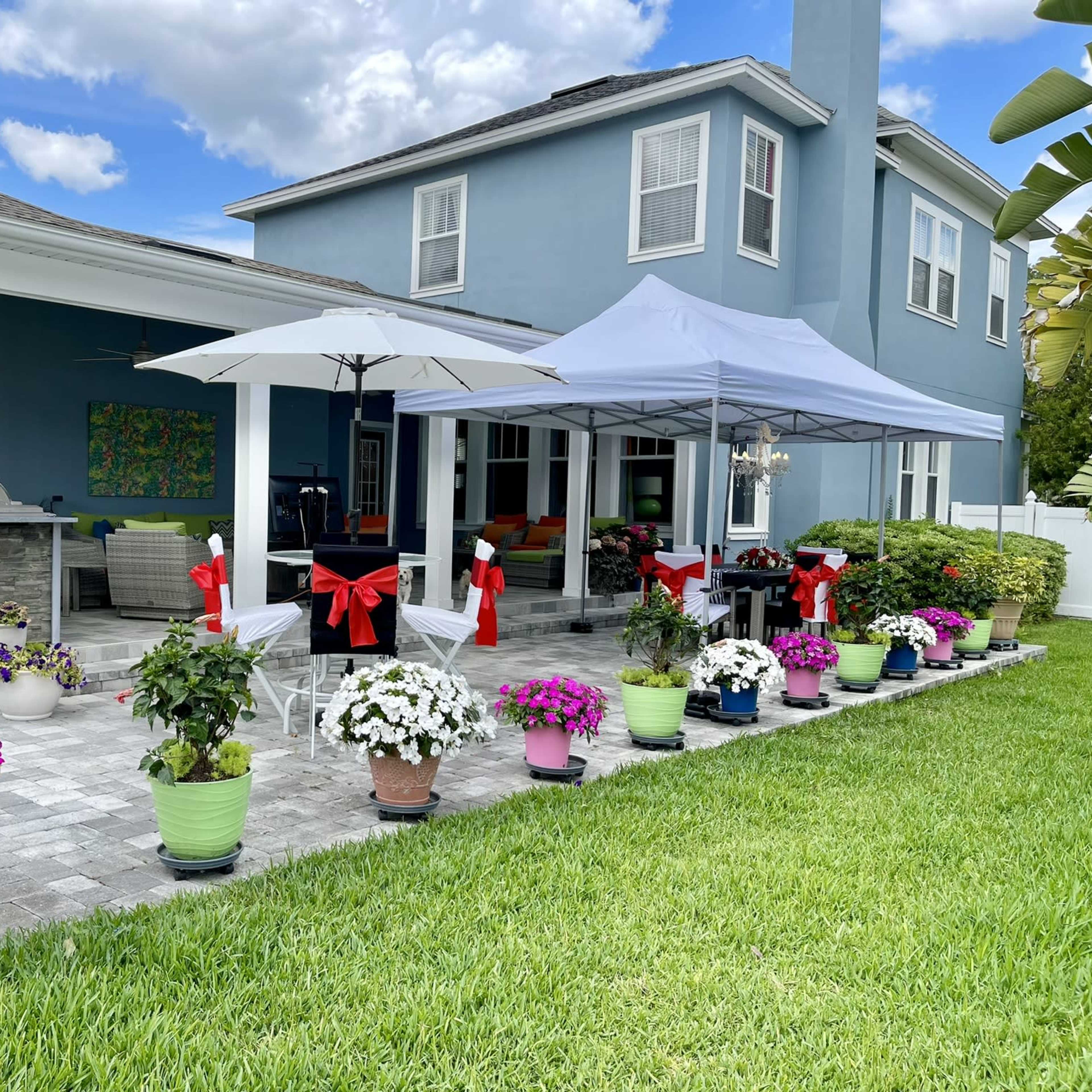 A covered patio area with colorful flower pots arranged along the edge, and two large umbrellas providing shade, against a backdrop of a blue house.