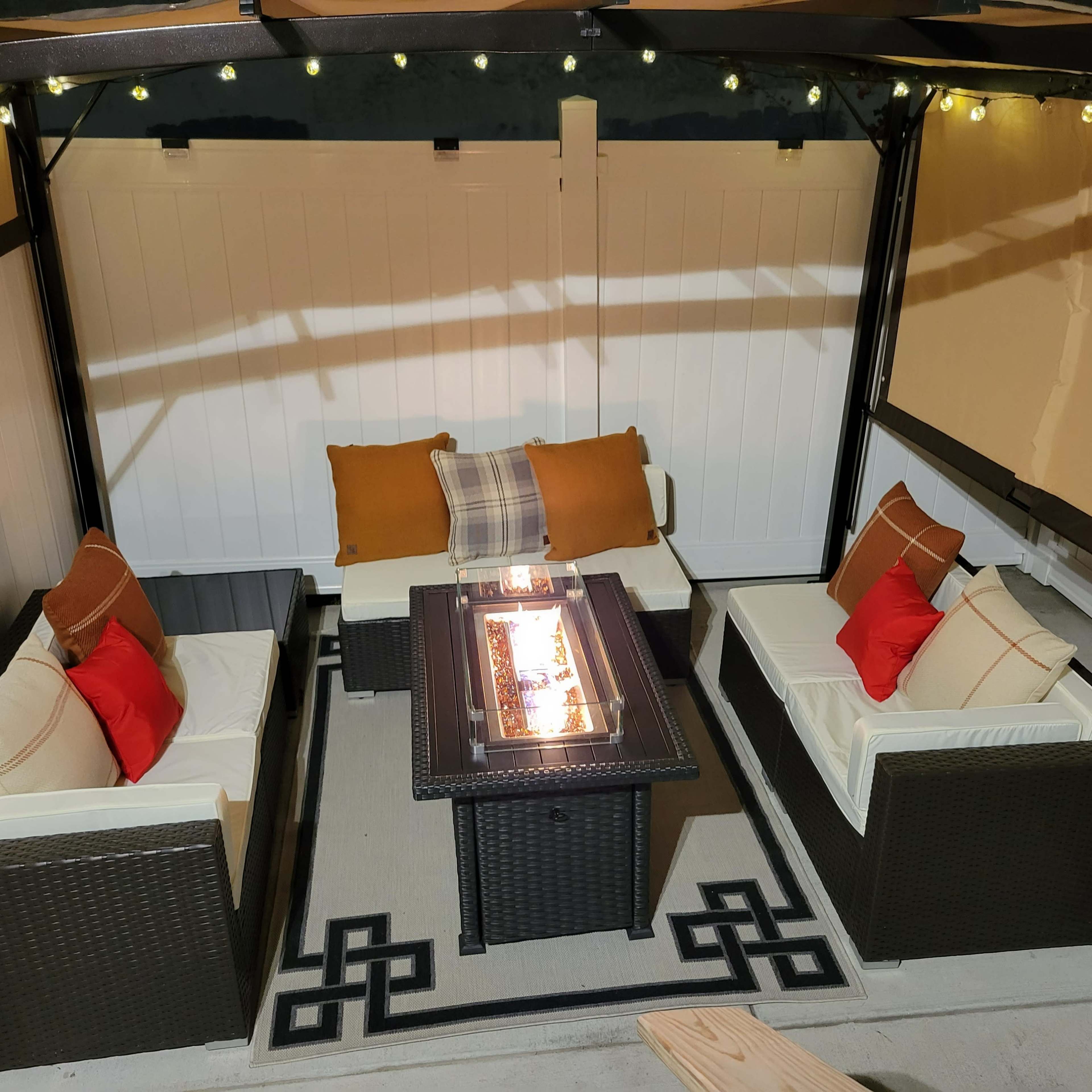 A seating area features a rectangular fire table surrounded by couches with cushions, all enclosed in a lighted gazebo.