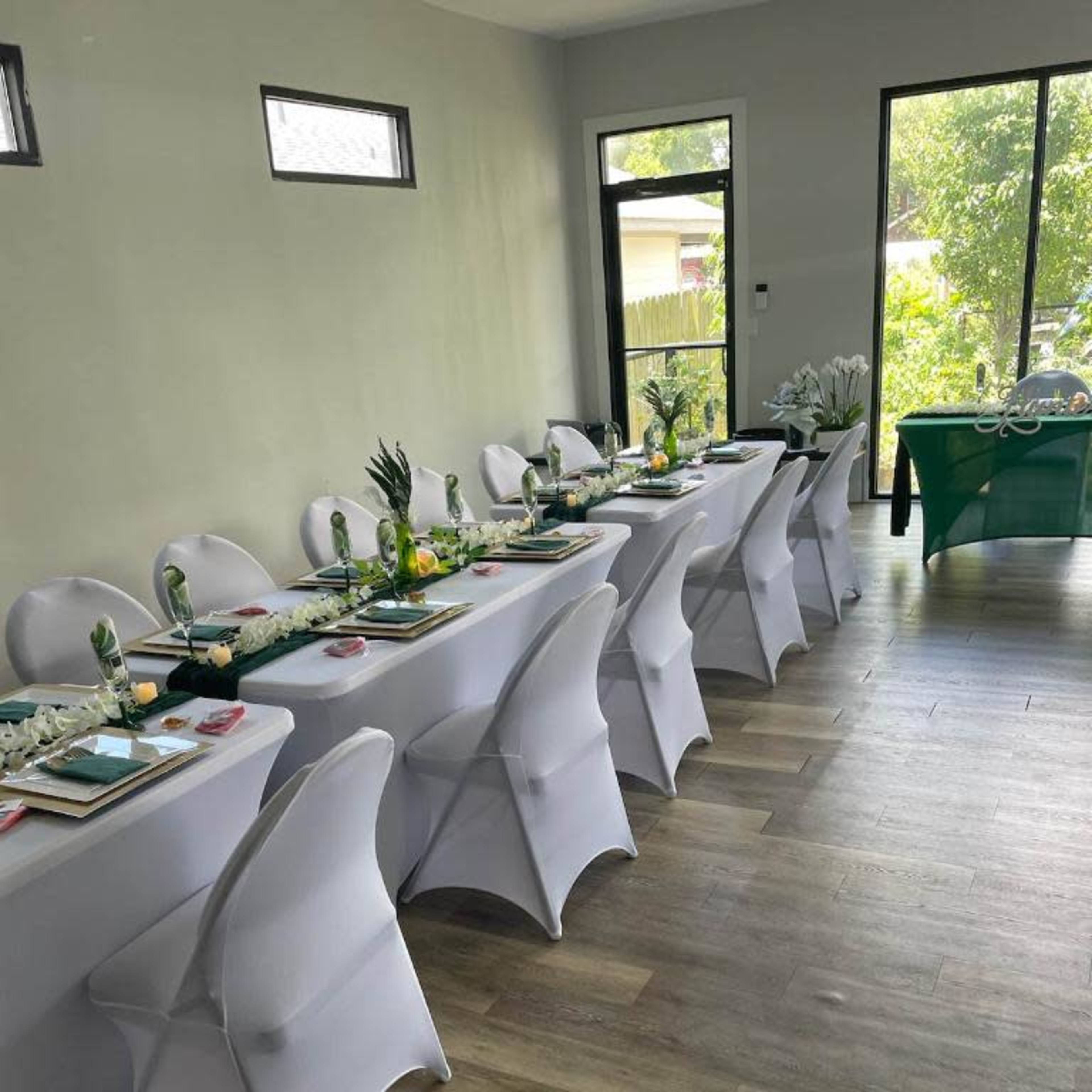 A long table is set with white tablecloths and decorations in a well-lit room, surrounded by white chairs and greenery outside the windows.