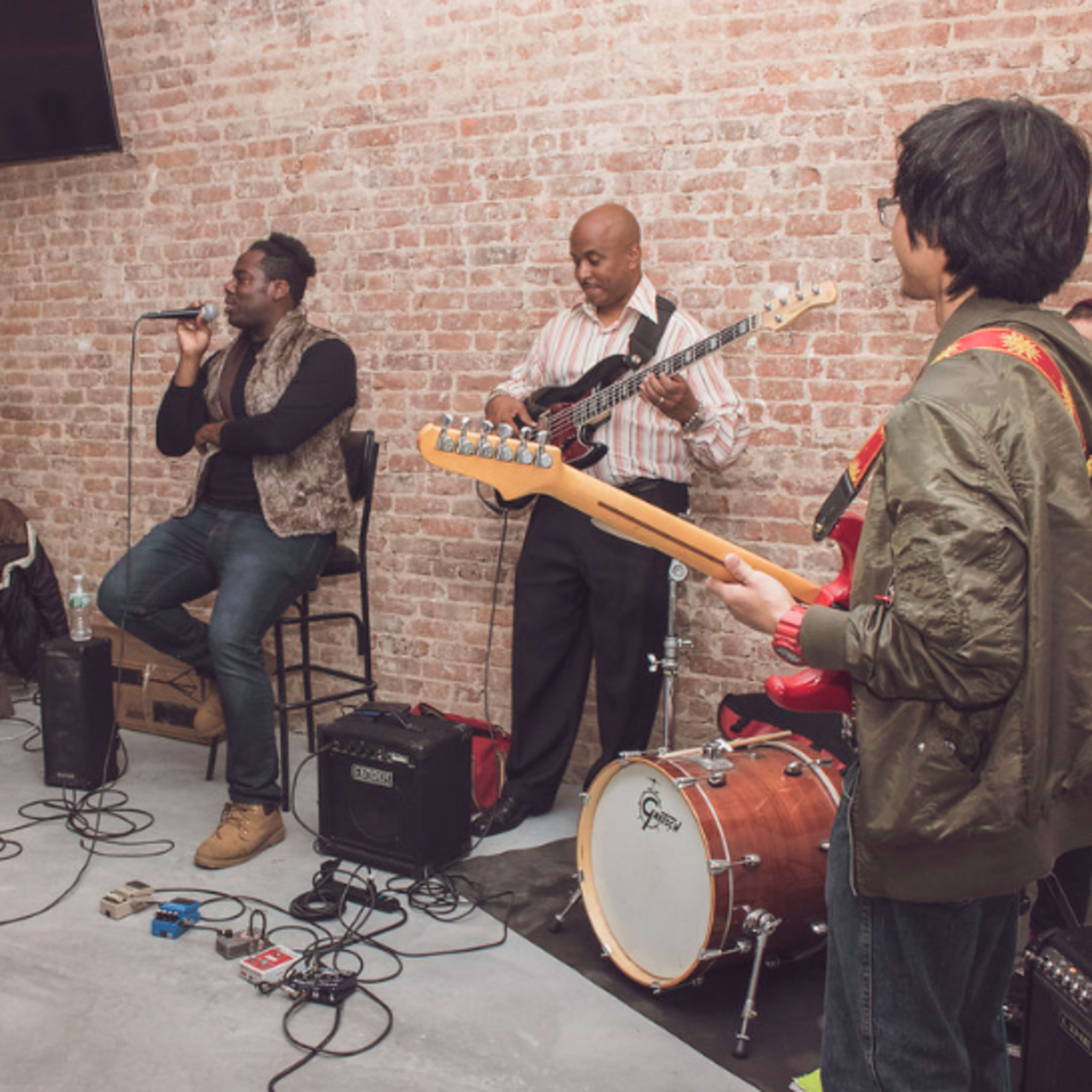 A group of musicians performs live music in a brick-walled venue, with one vocalist and three instrumentalists playing guitar and drums.