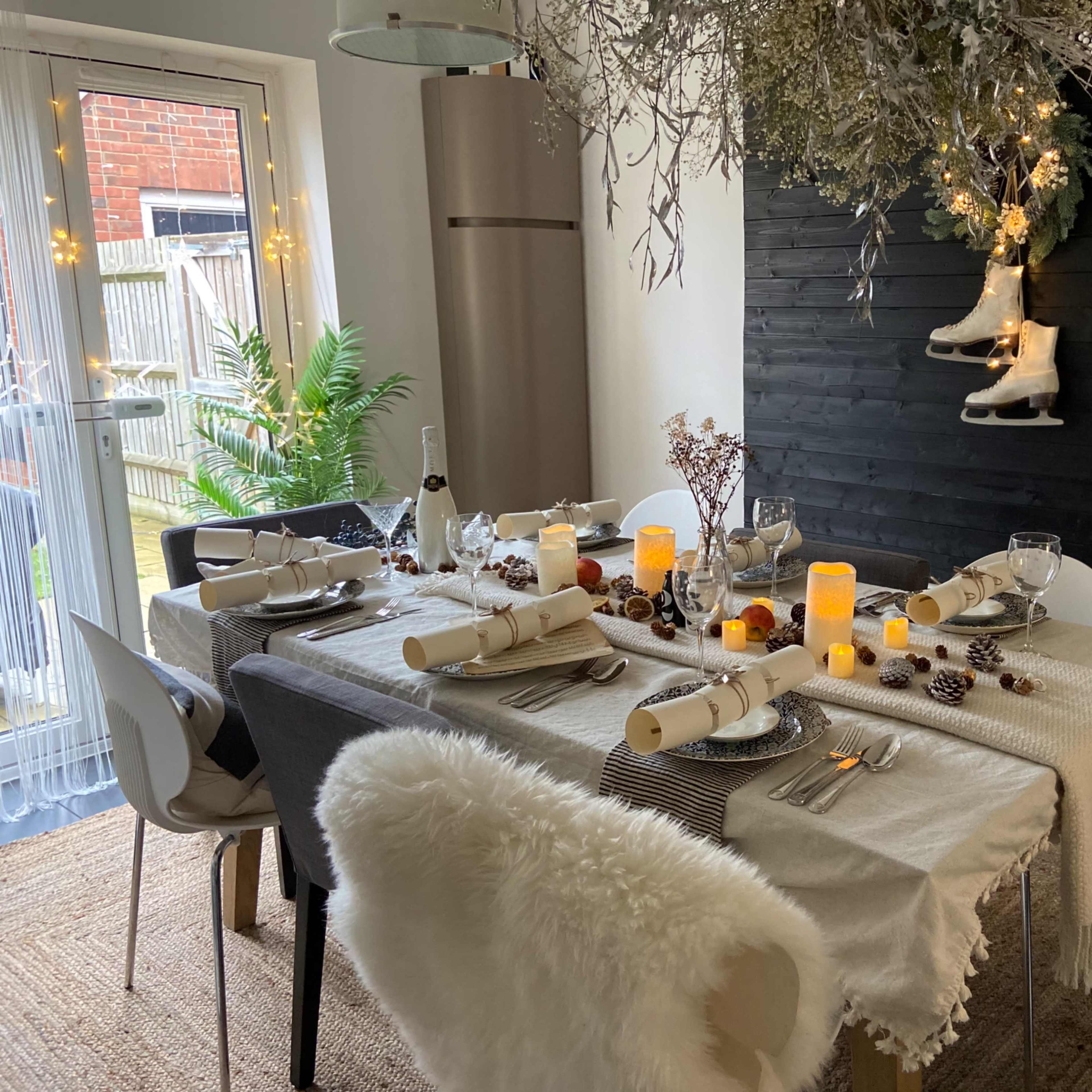 A dining table is set with elegant tableware and decorations, featuring flickering candles, pinecones, and hanging ice skates above.