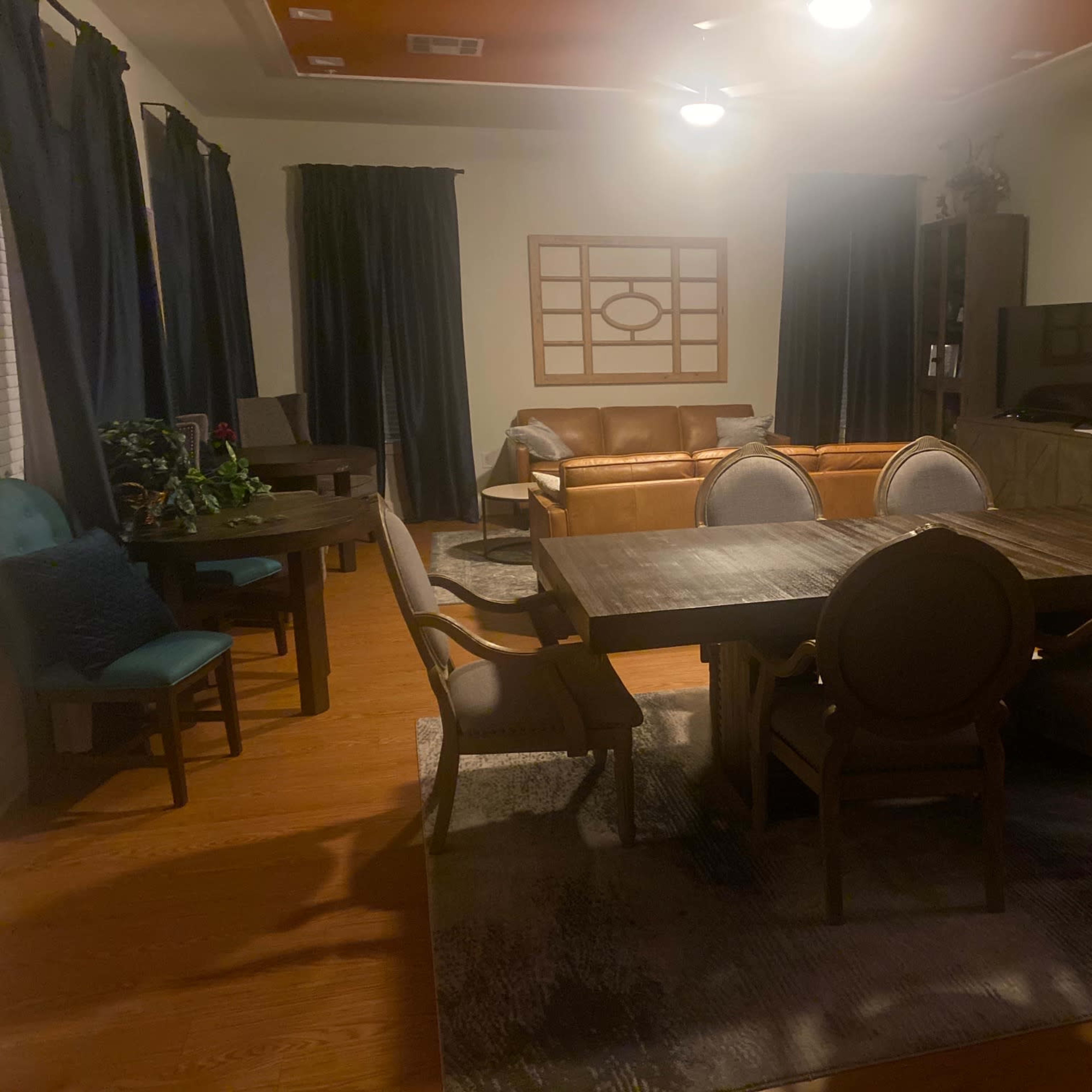 A living room features a large dining table with chairs, a seating area with a sofa, and shelves against the walls.