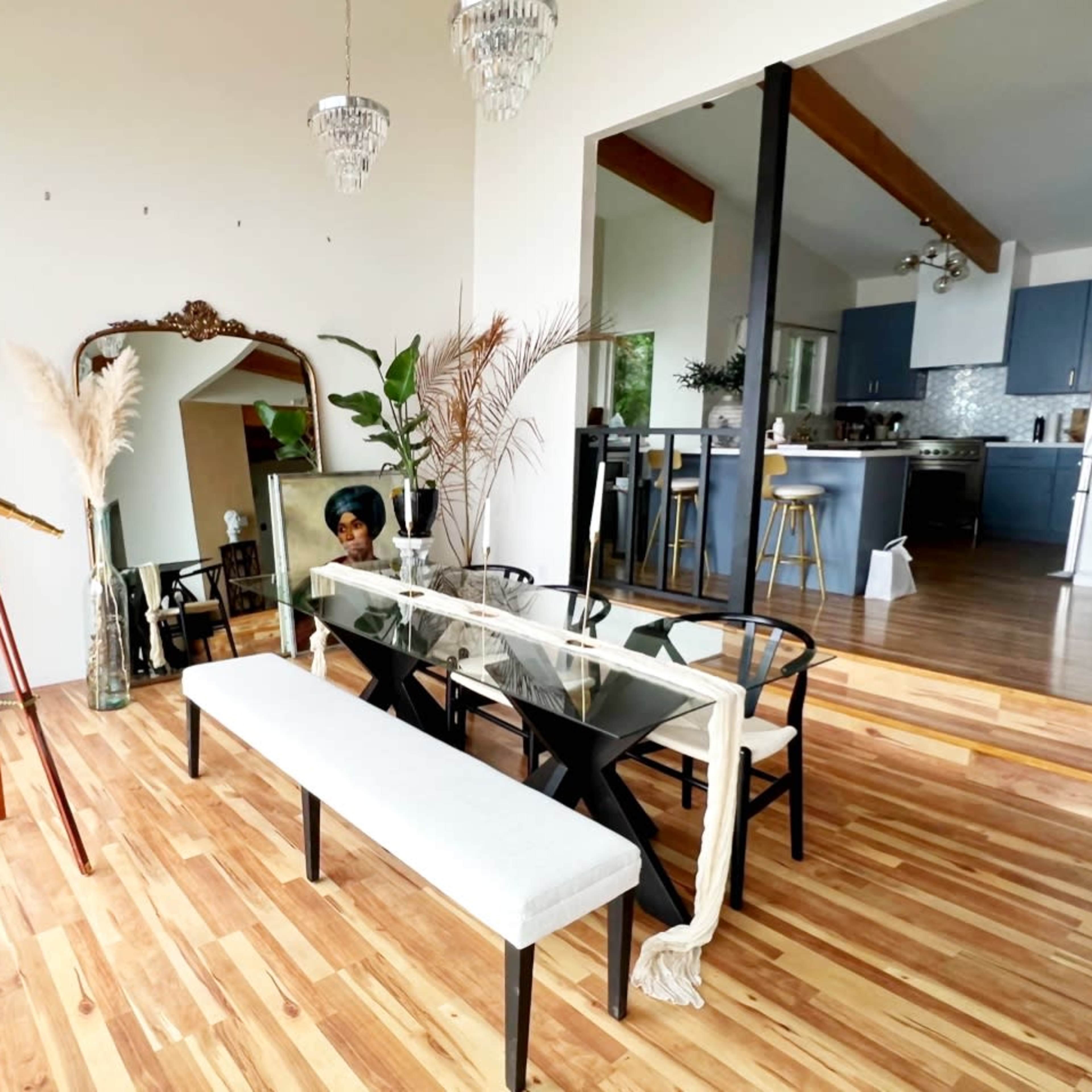 A modern dining area features a glass table, black chairs, a wooden bench, and decor elements including a large mirror, a telescope, and potted plants.