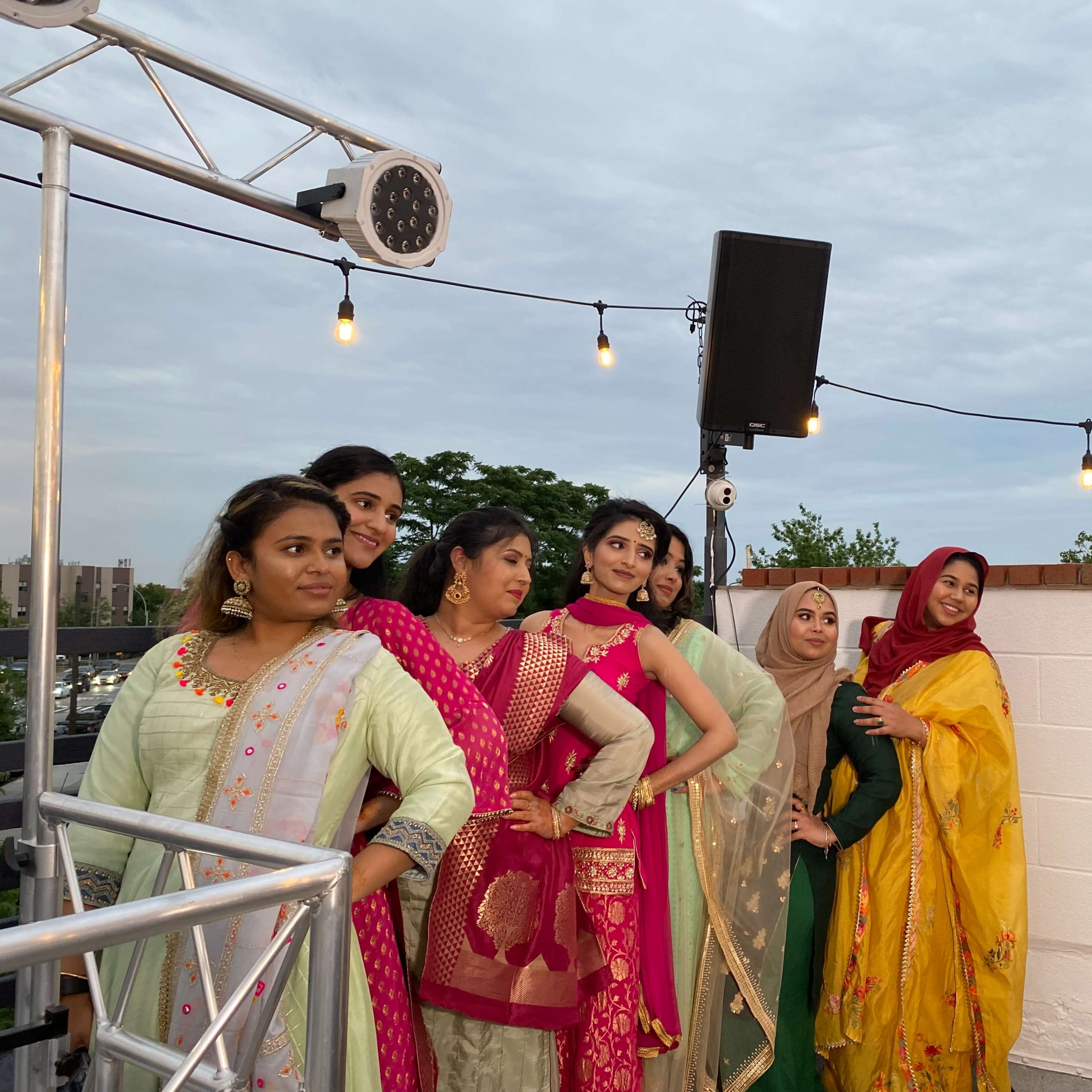 A group of women, dressed in traditional attire, poses for a photo on a rooftop with string lights and a city view in the background.