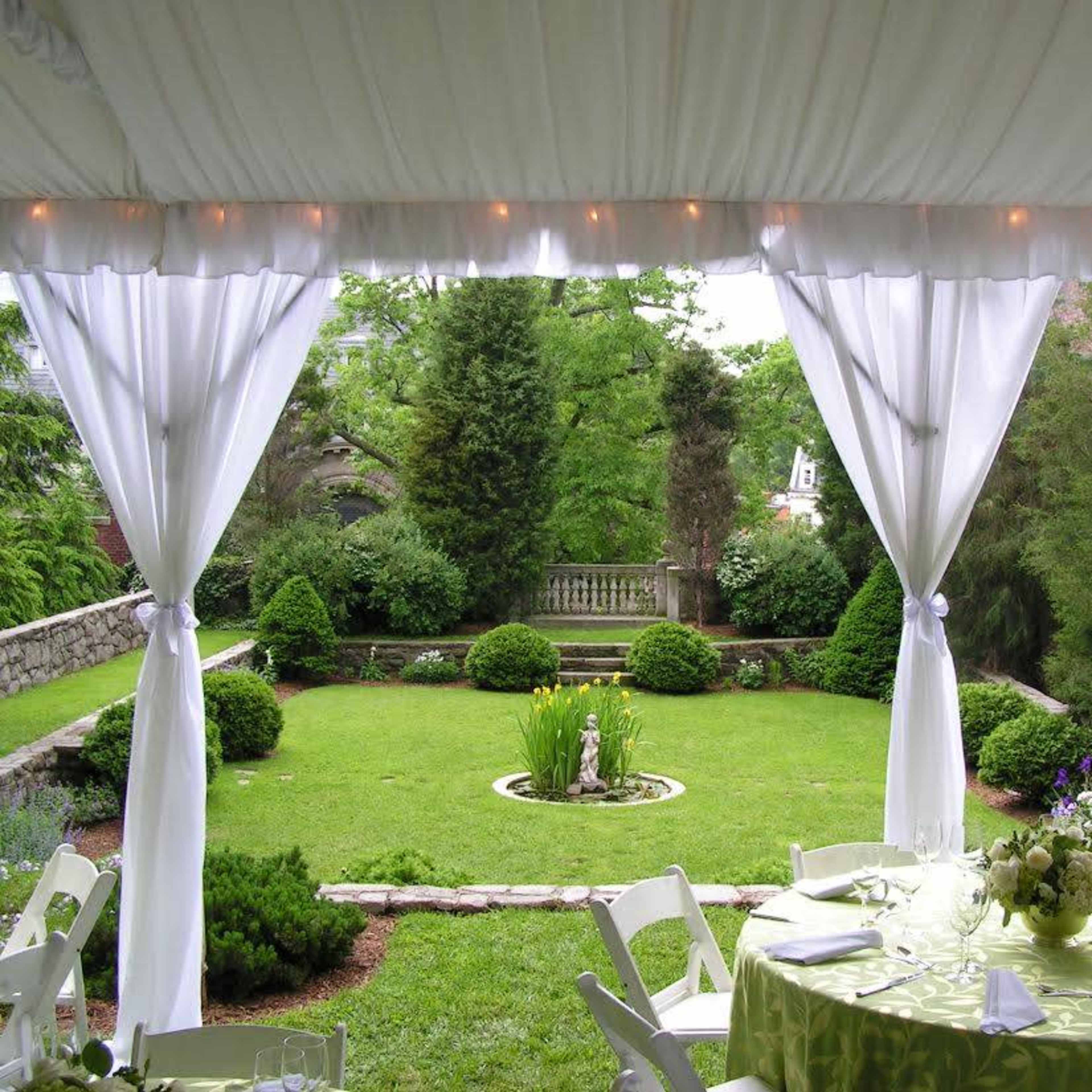 A white tent with draped fabric overlooking a well-maintained garden featuring greenery and a central fountain.