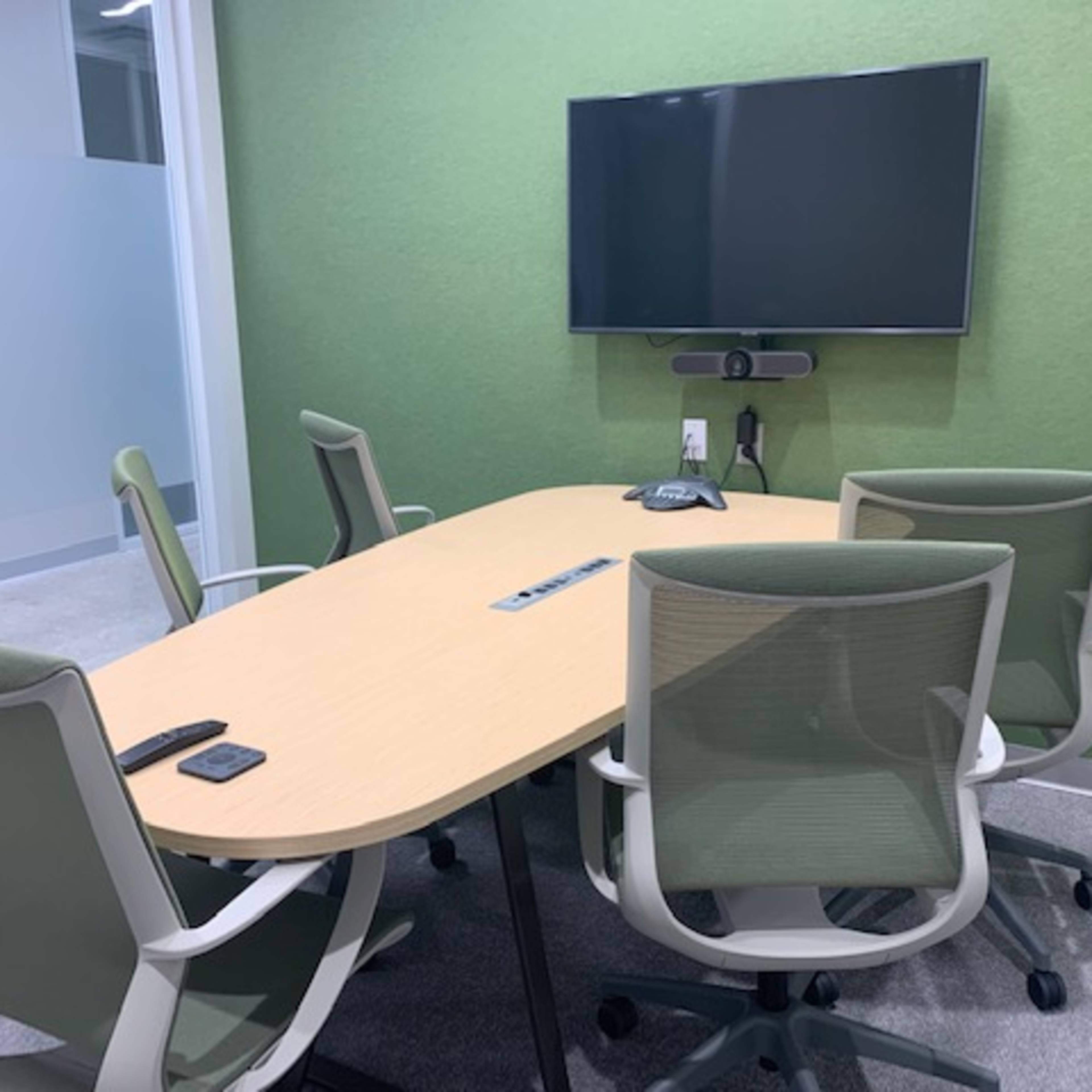 A conference room features a long wooden table surrounded by four light green chairs and a wall-mounted television.
