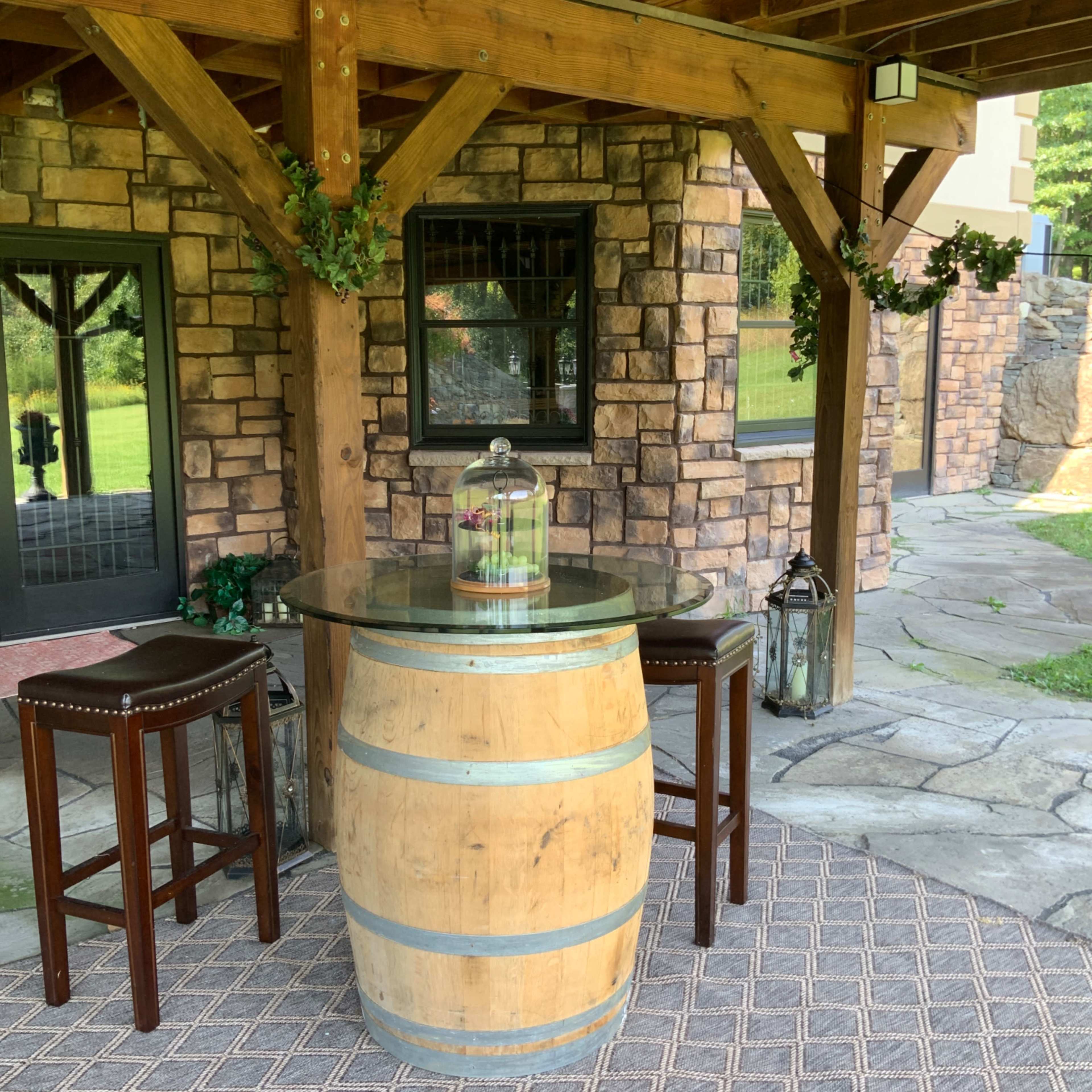 A wooden barrel serves as a table, topped with a glass surface, accompanied by two high-backed chairs under a wooden structure with stone walls.