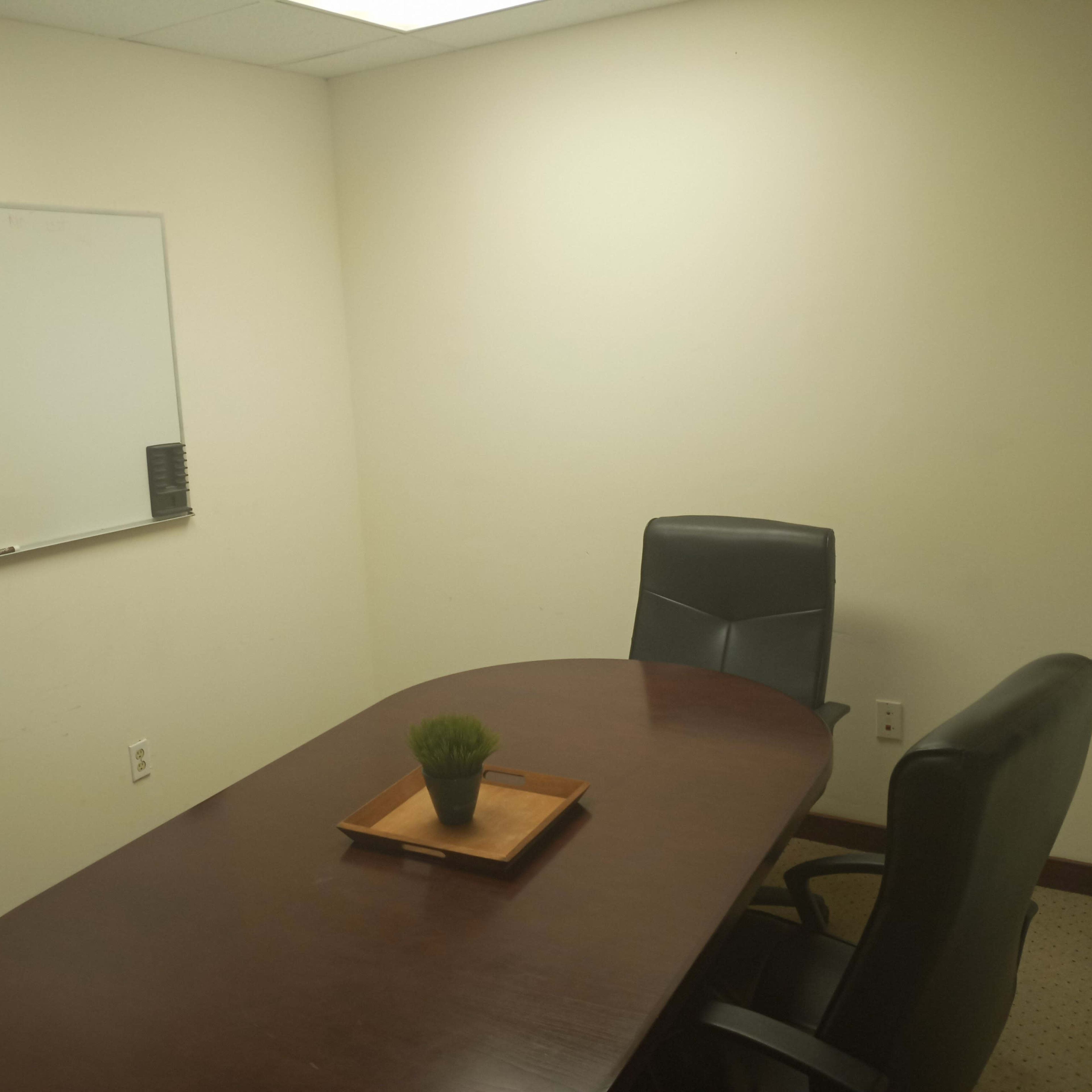 A small conference room features a wooden table with a plant centerpiece and two black chairs, alongside a whiteboard displaying handwritten notes.