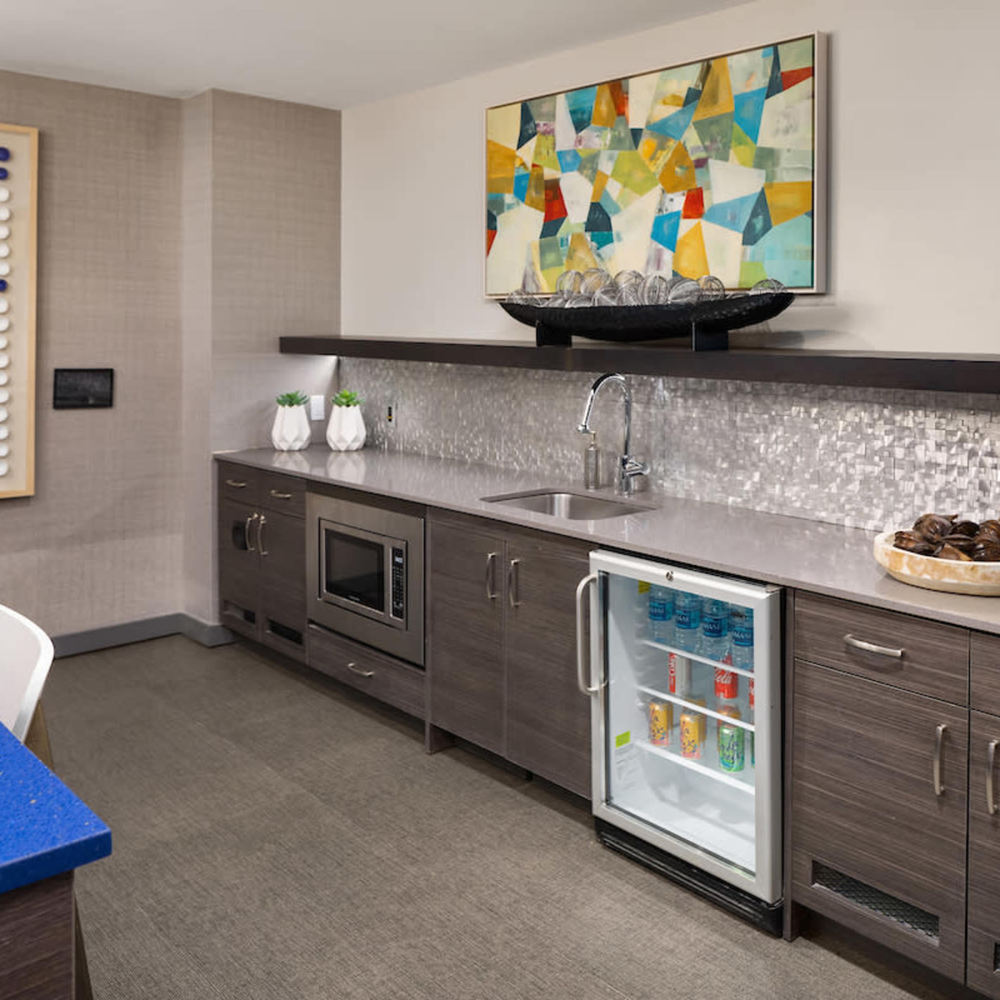 A modern kitchen with dark wood cabinetry, a stainless steel sink, and a small refrigerator displaying beverages, alongside a counter topped with decorative items and a colorful abstract painting above.