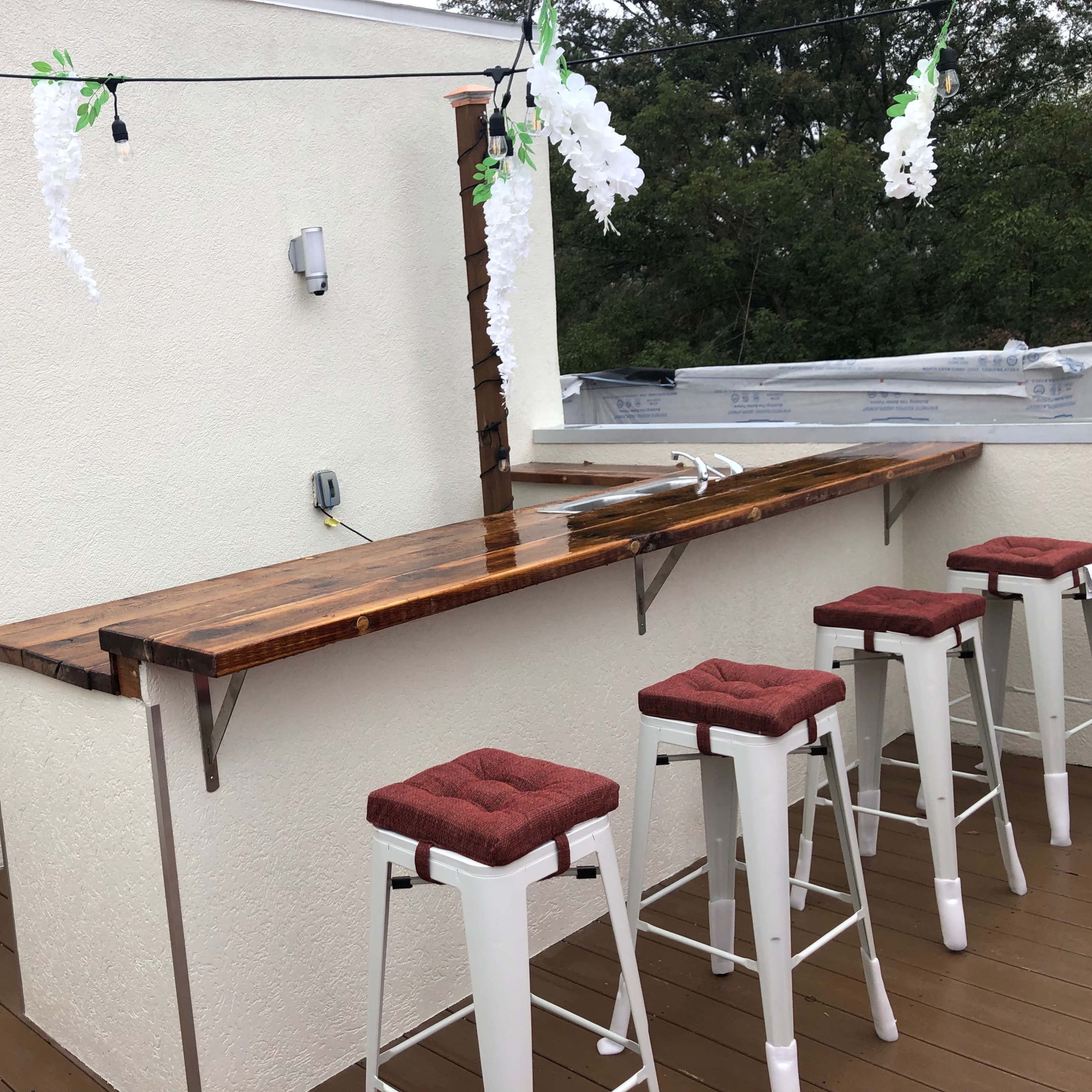 A rooftop bar with a wooden countertop and four white stools, decorated with hanging plants and lights.
