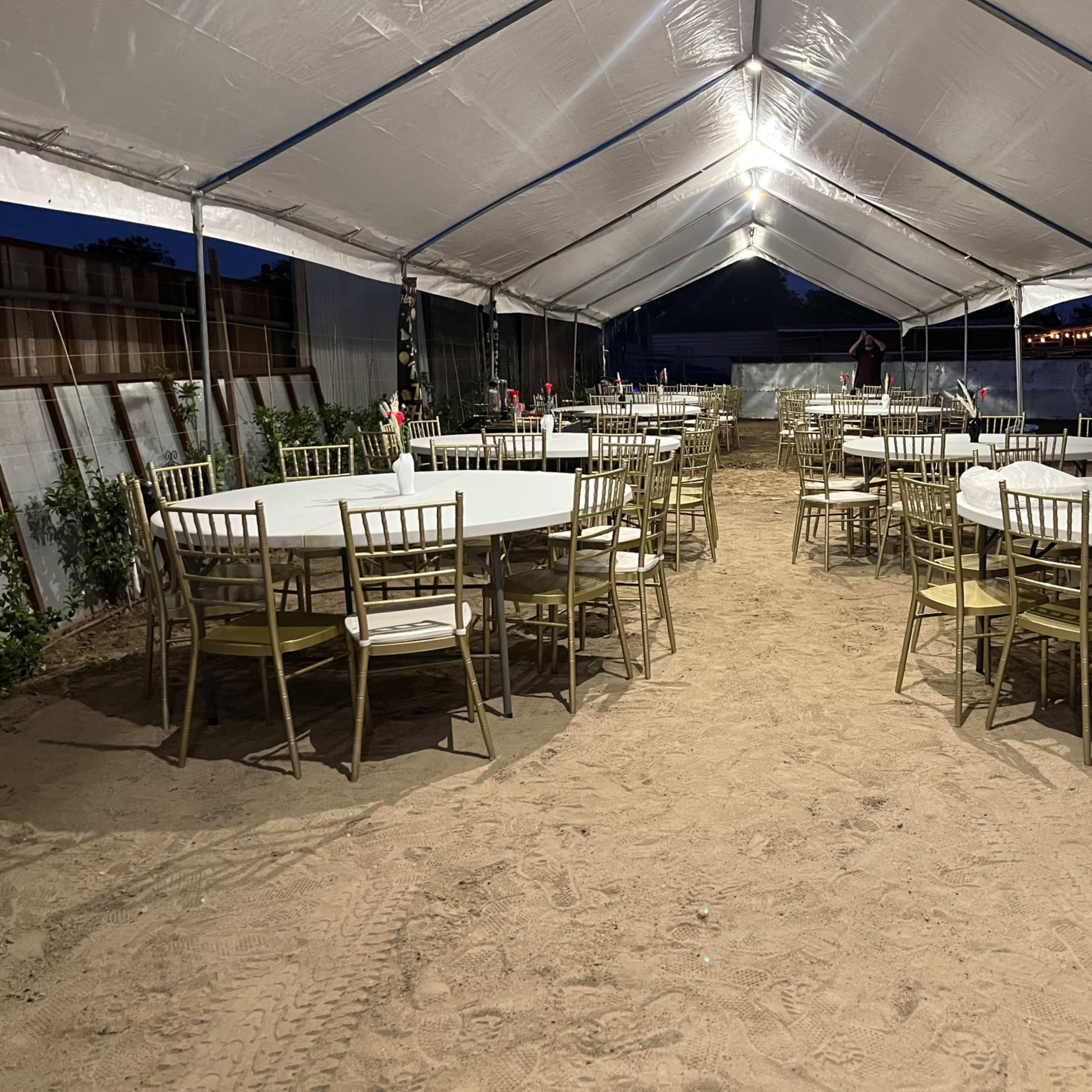 A large, open tent is set up with circular tables and chairs arranged on sandy ground, illuminated by overhead lights.