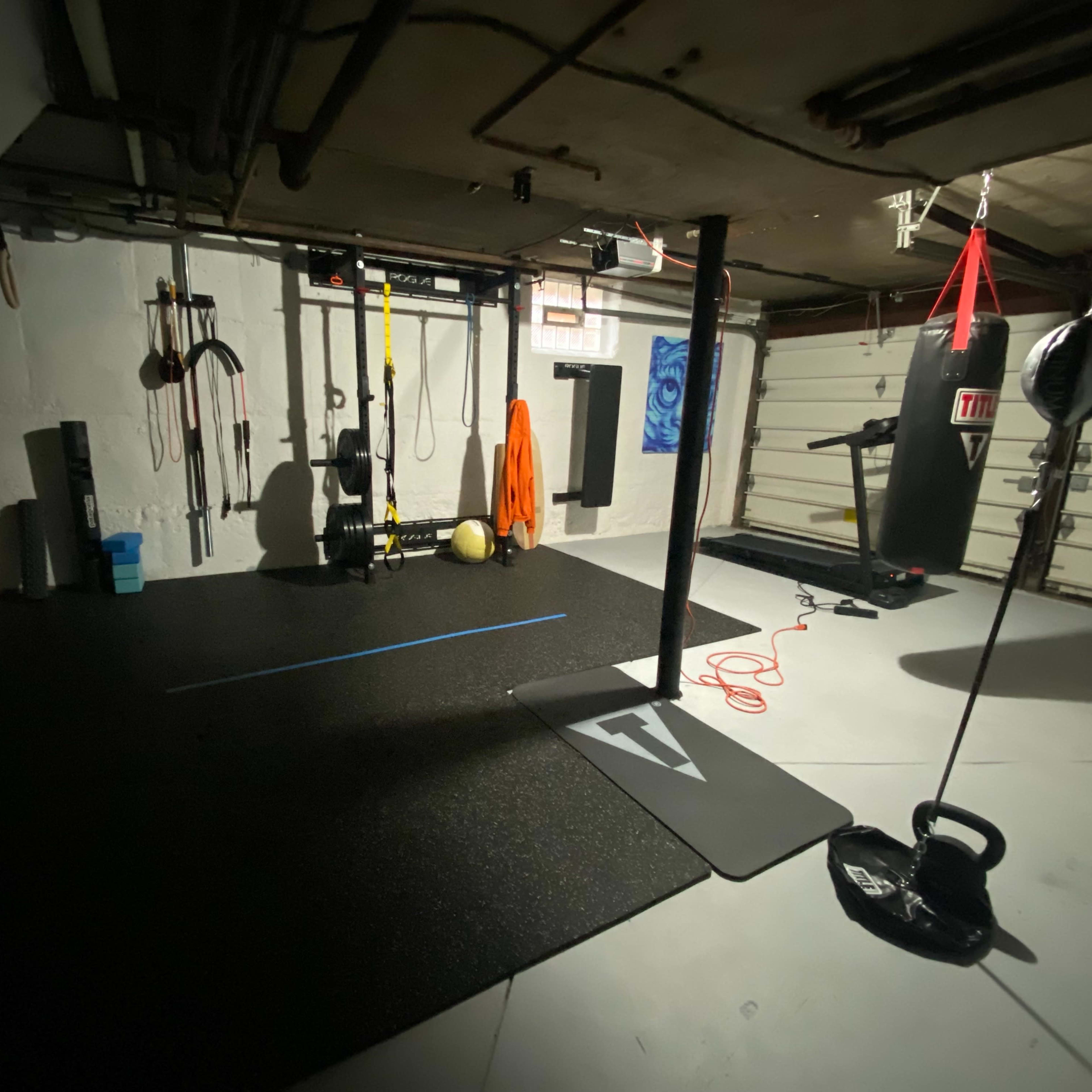 A dimly lit home gym featuring a punching bag, kettlebell, exercise mats, and various workout equipment hanging on the walls.