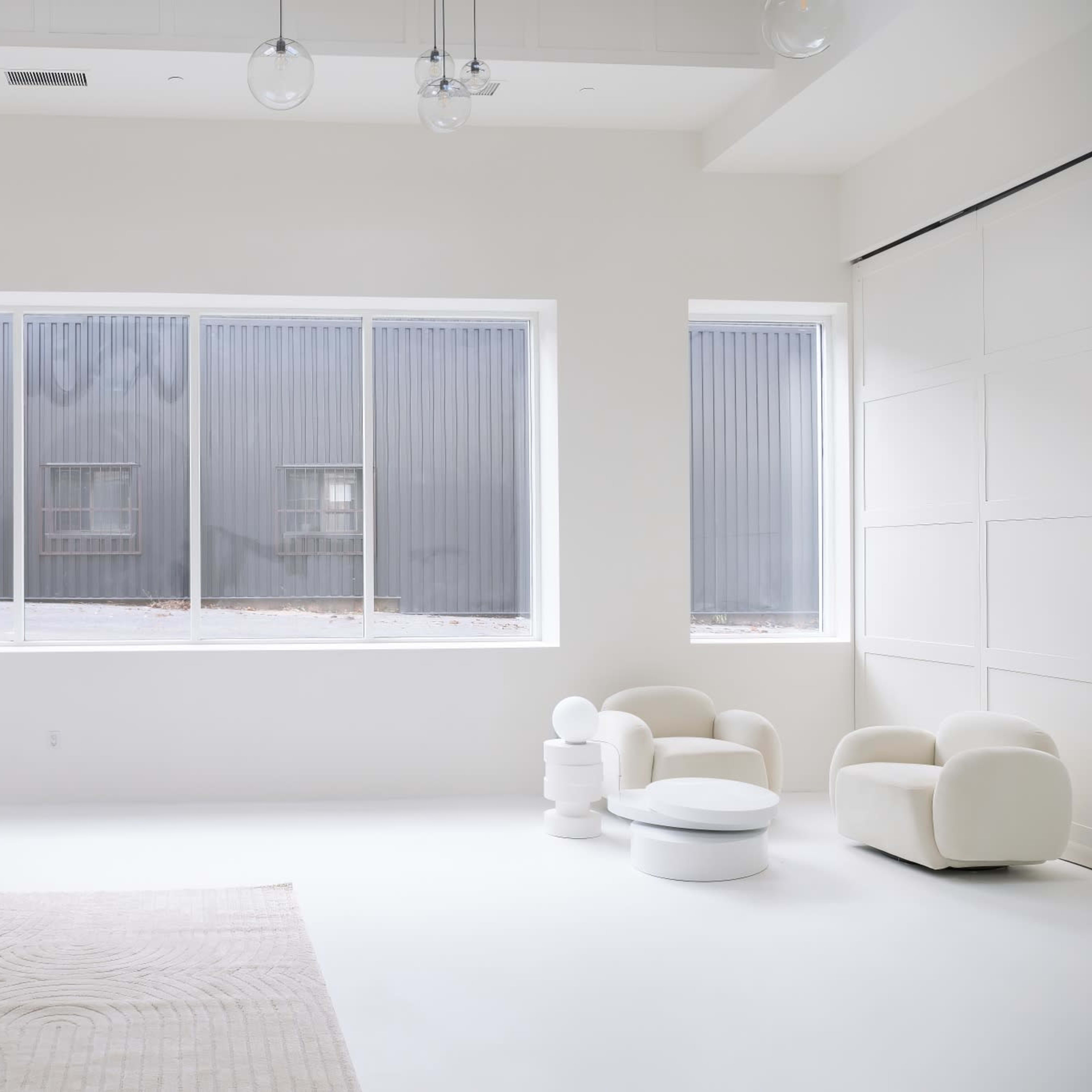 A minimalist, white interior space featuring a sofa, round coffee table, and modern chairs, with large windows revealing a gray exterior wall.
