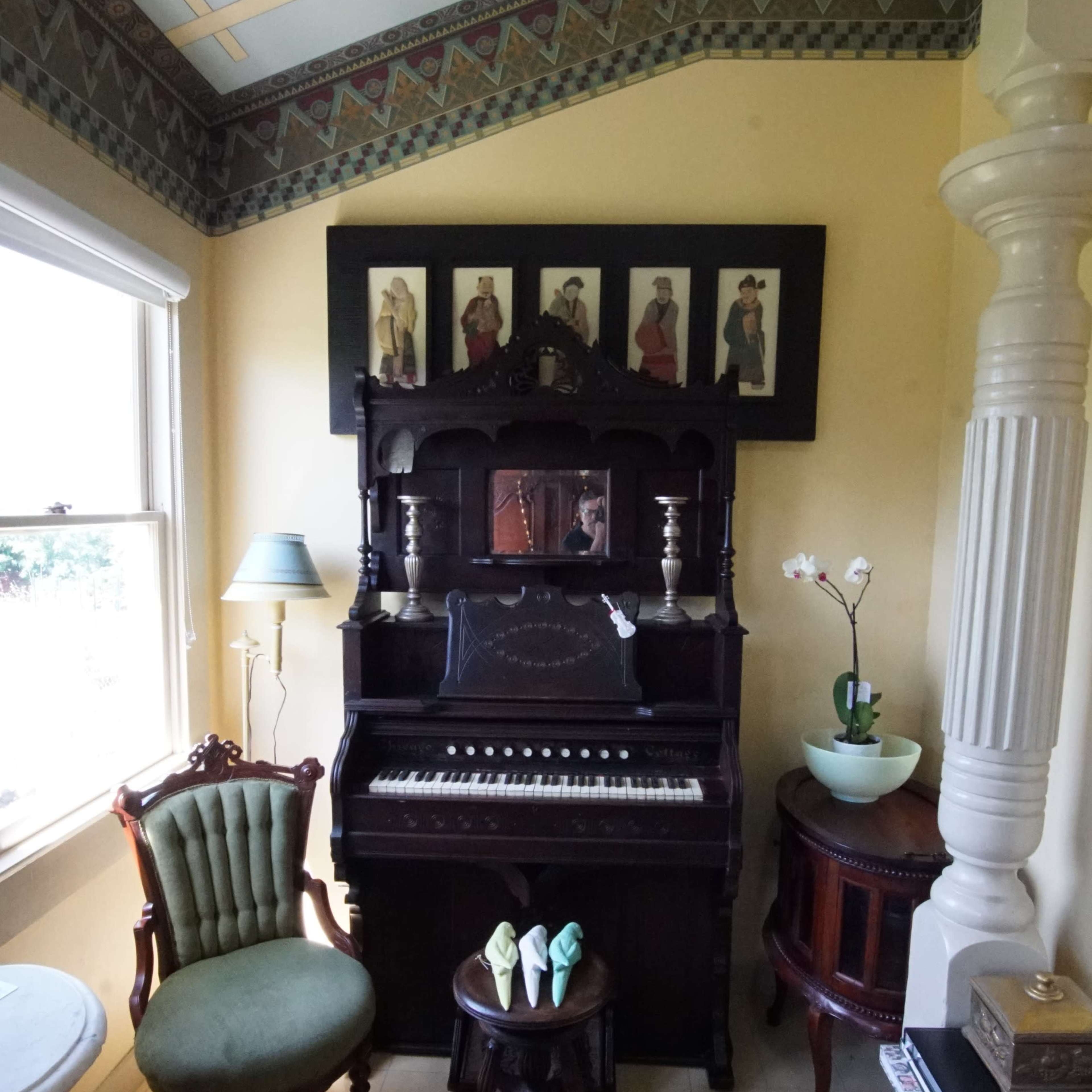 A vintage piano positioned between a green upholstered chair and a wooden side table in a sunlit room with ornate ceiling details.