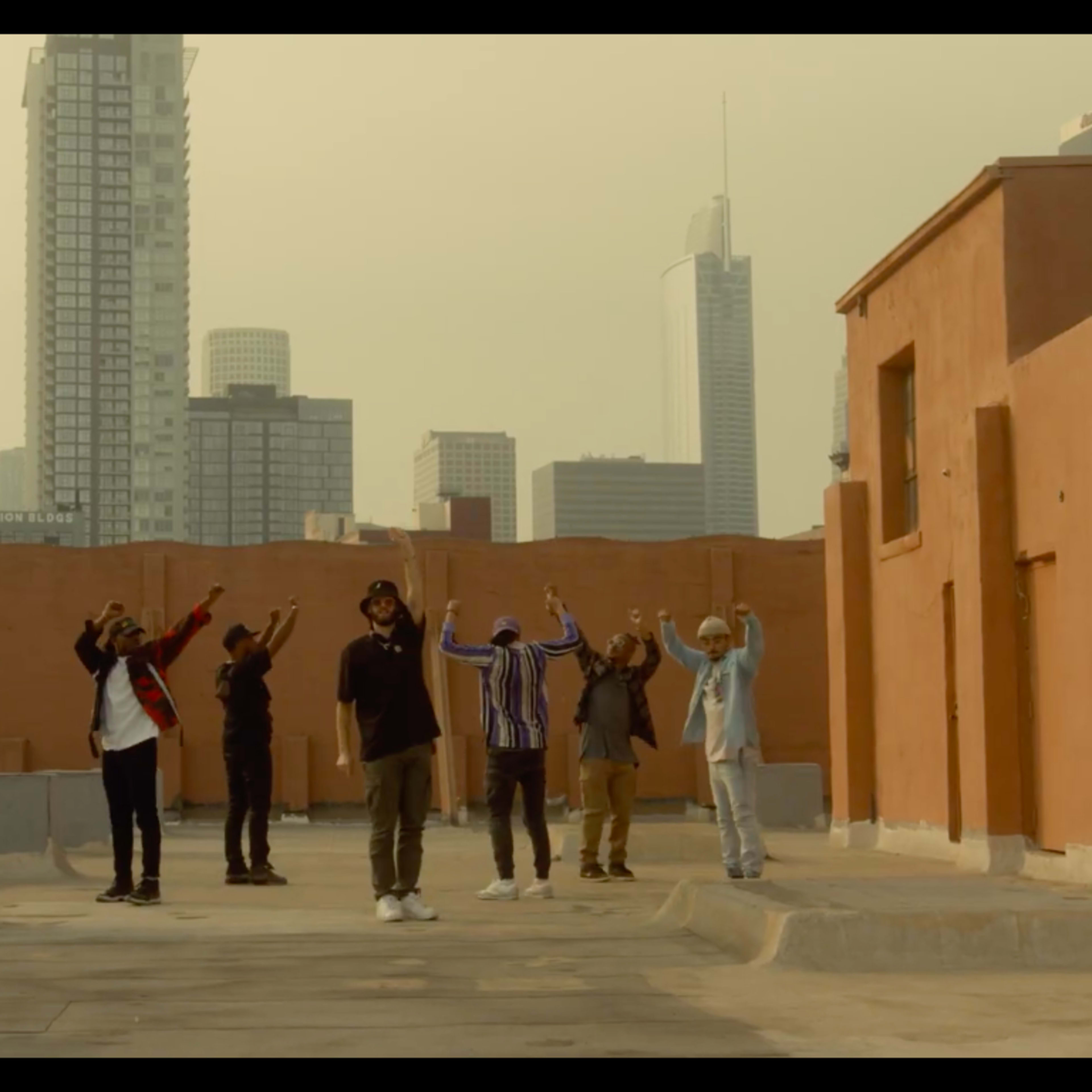 A group of seven people stands on a rooftop with their arms raised, featuring a skyline of tall buildings in the background.