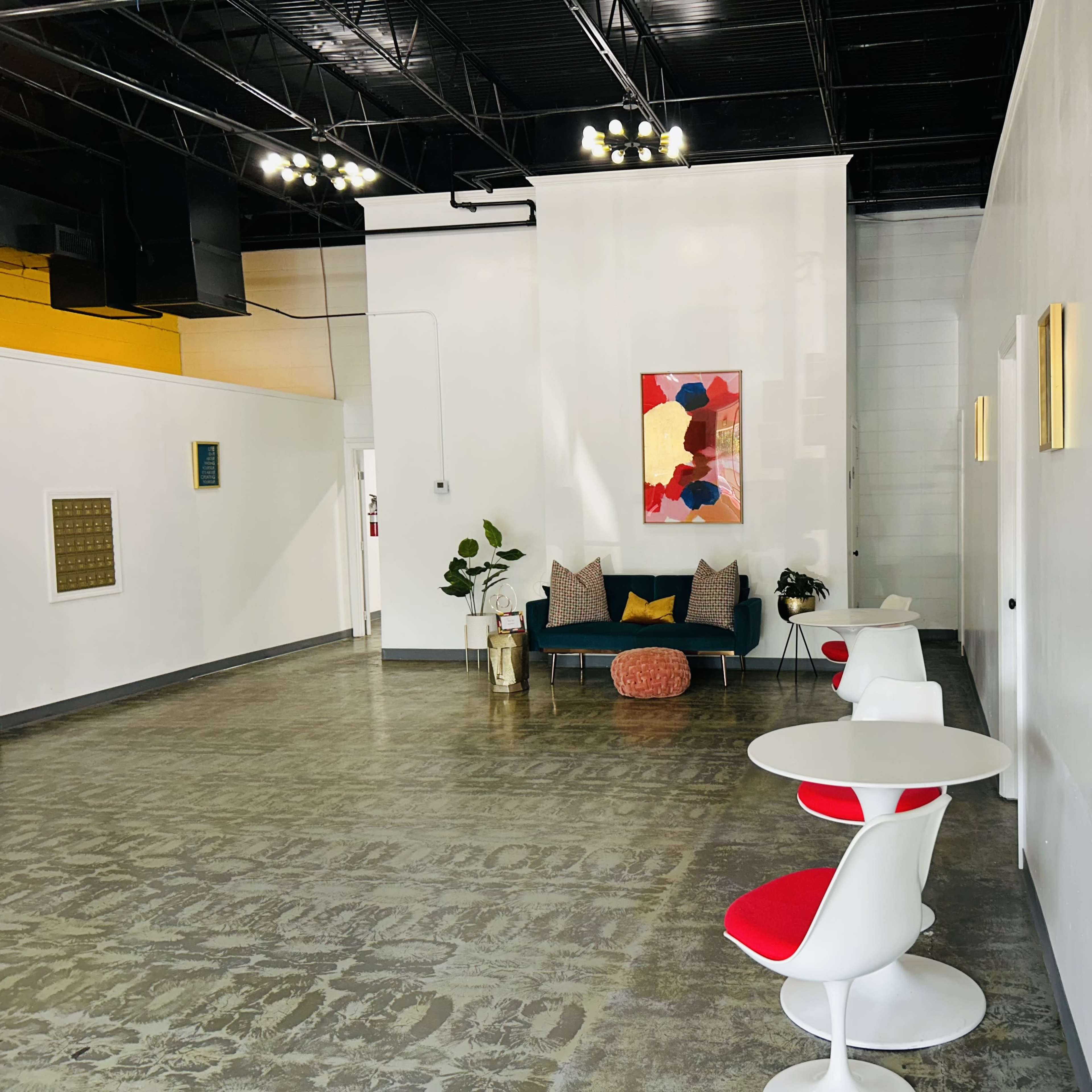 A spacious, modern interior with white walls, a colorful abstract painting, a blue couch, and minimalist round tables with red accents.