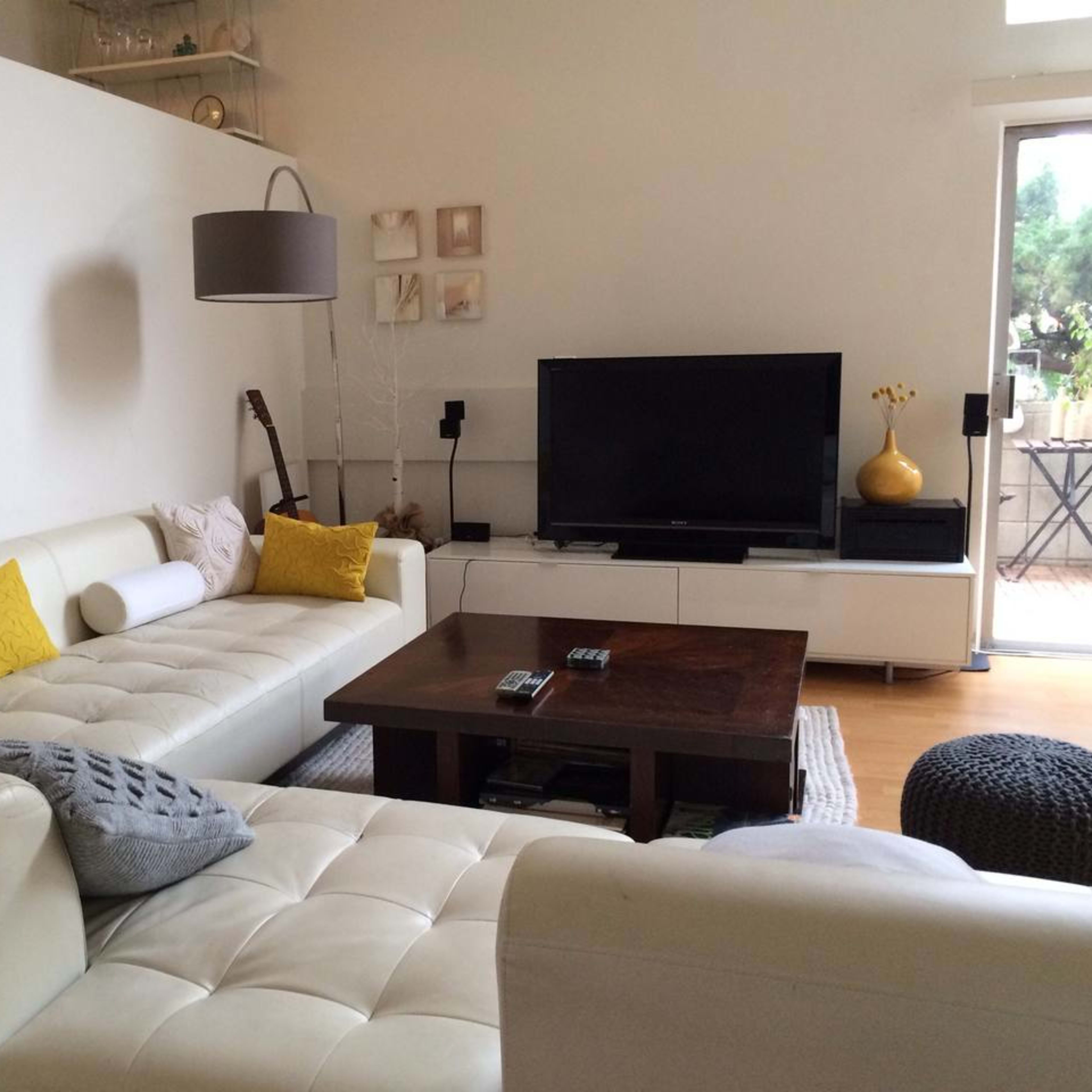 A modern living room features a white sectional sofa with yellow accent pillows, a wooden coffee table, and a large television against a light-colored wall.
