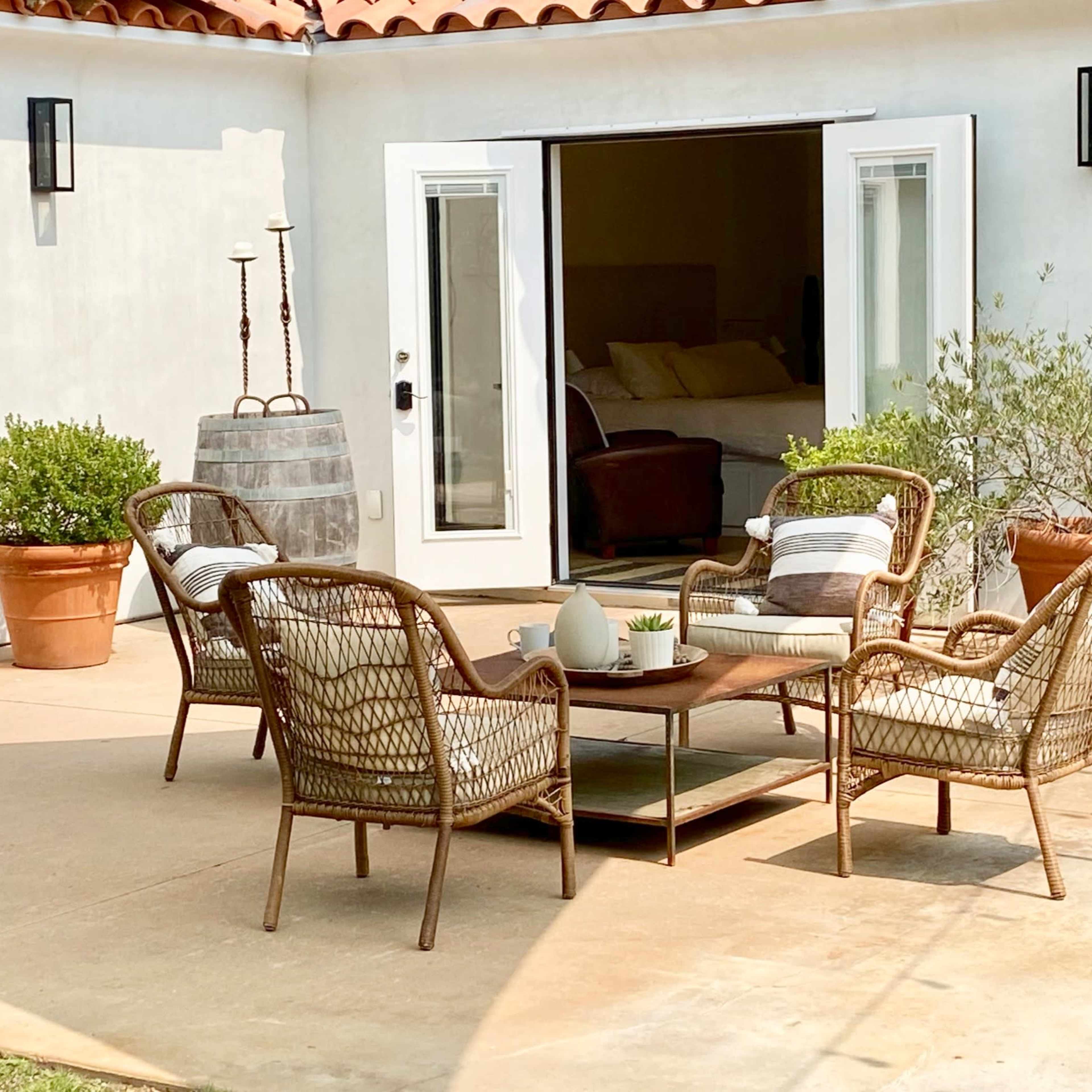 A patio area features a table with four wicker chairs surrounded by potted plants and accessed by two open doors.