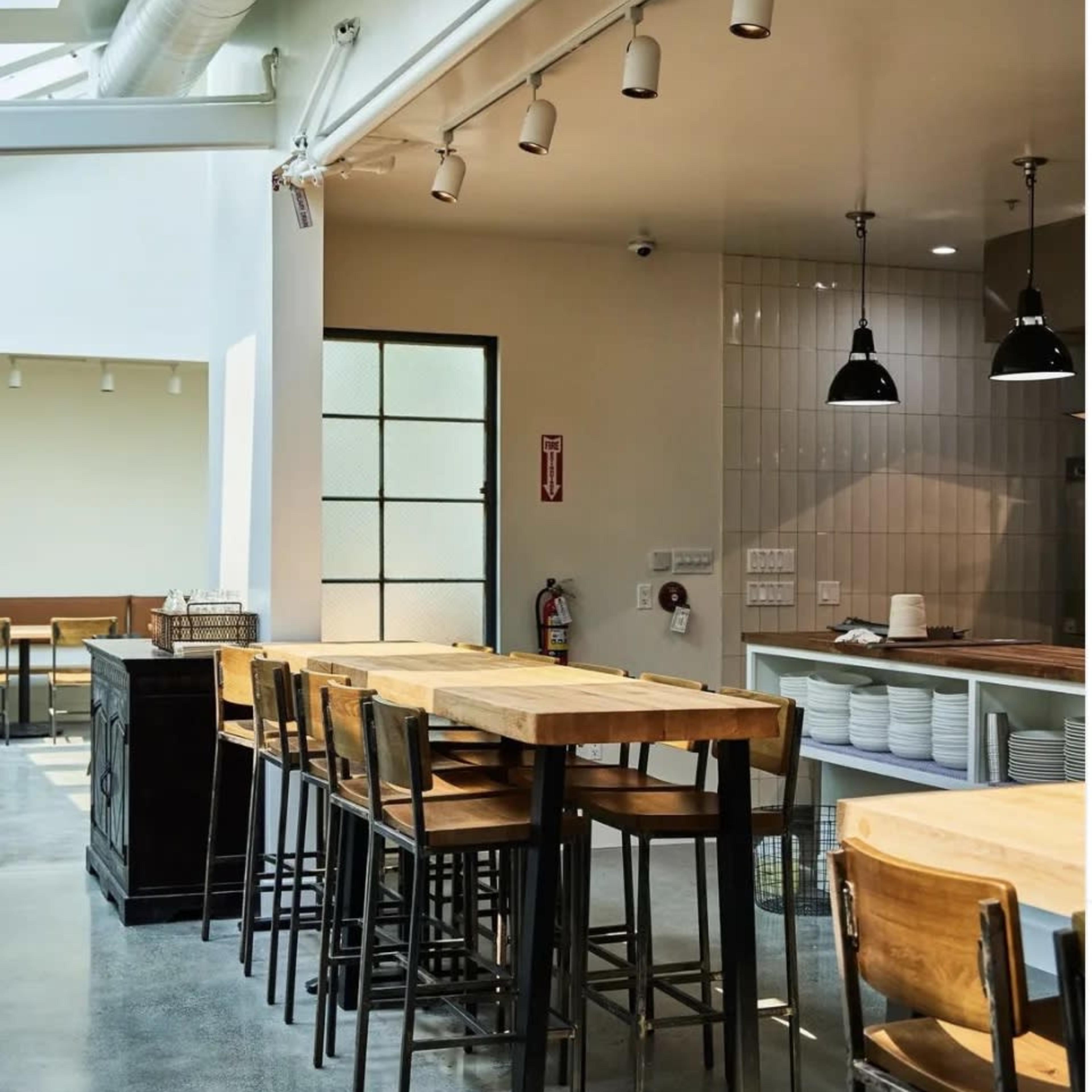 A modern cafe with wooden tables and high stools, featuring an open kitchen and large windows allowing natural light to enter.