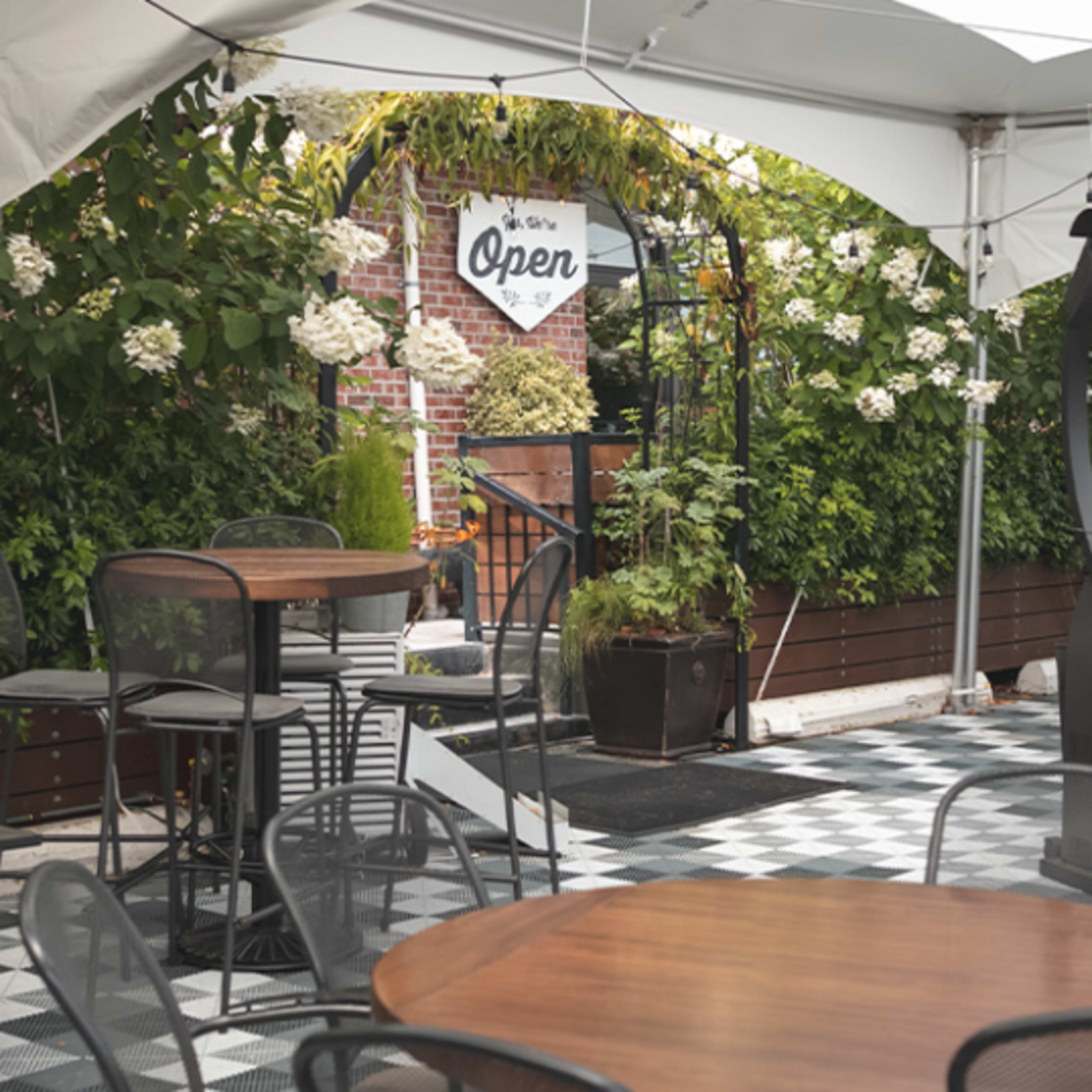 An outdoor seating area under a white tent, featuring several round tables and chairs, with lush greenery and a sign indicating that the space is open.