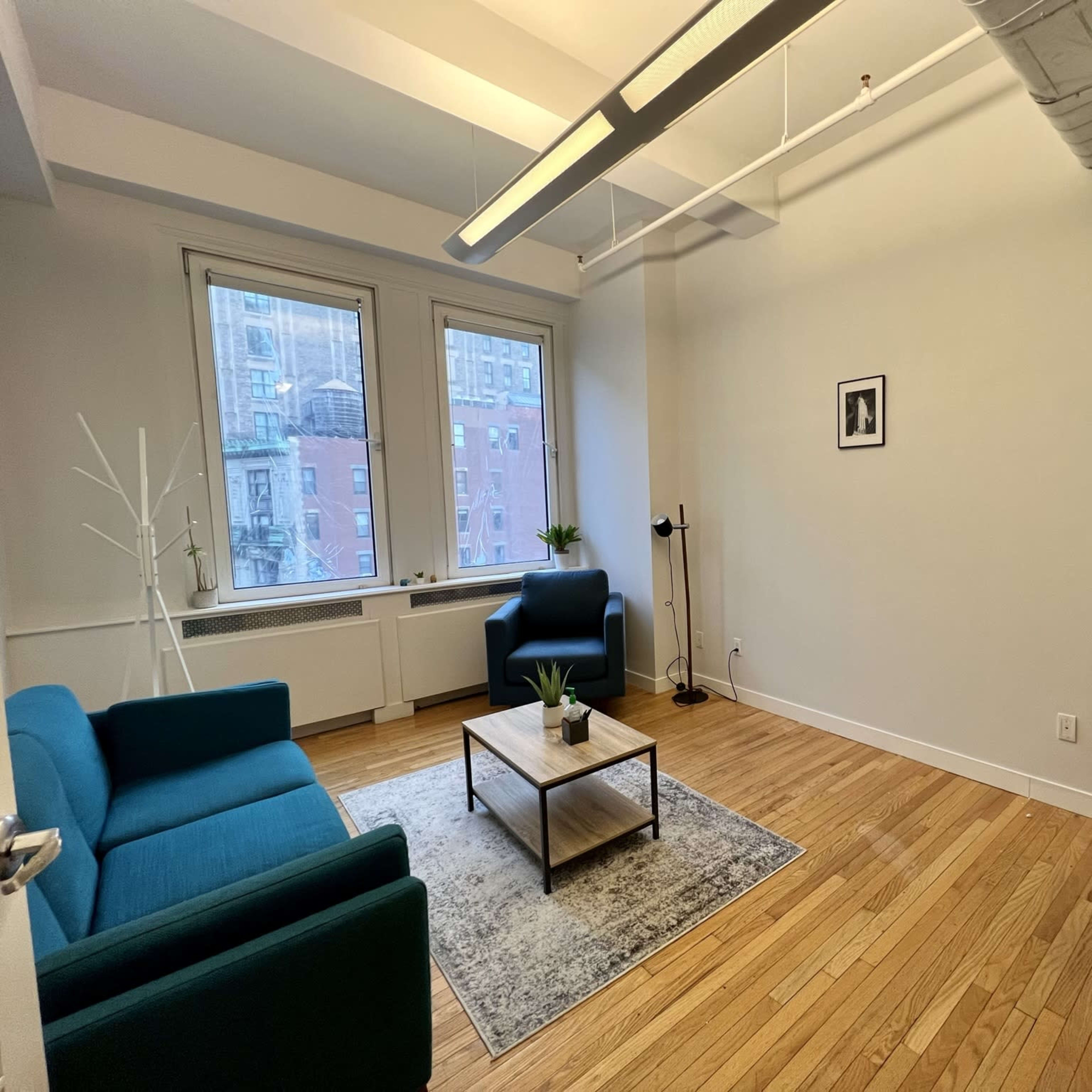 A minimalistic office space with two green chairs, a coffee table, and a desk in a well-lit room featuring large windows.