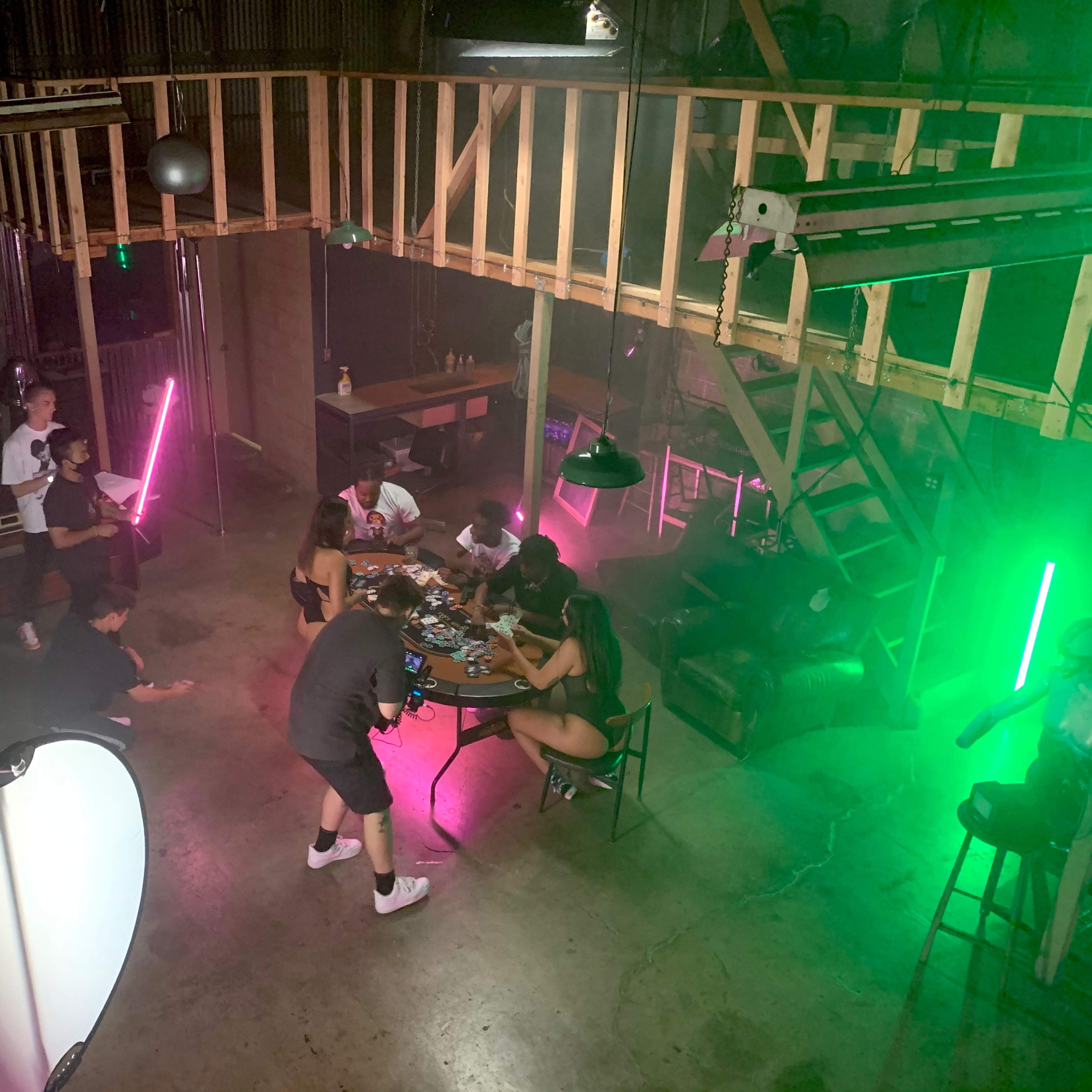 A group of people gathers around a table in a dimly lit industrial space, with colorful neon lights illuminating the scene.
