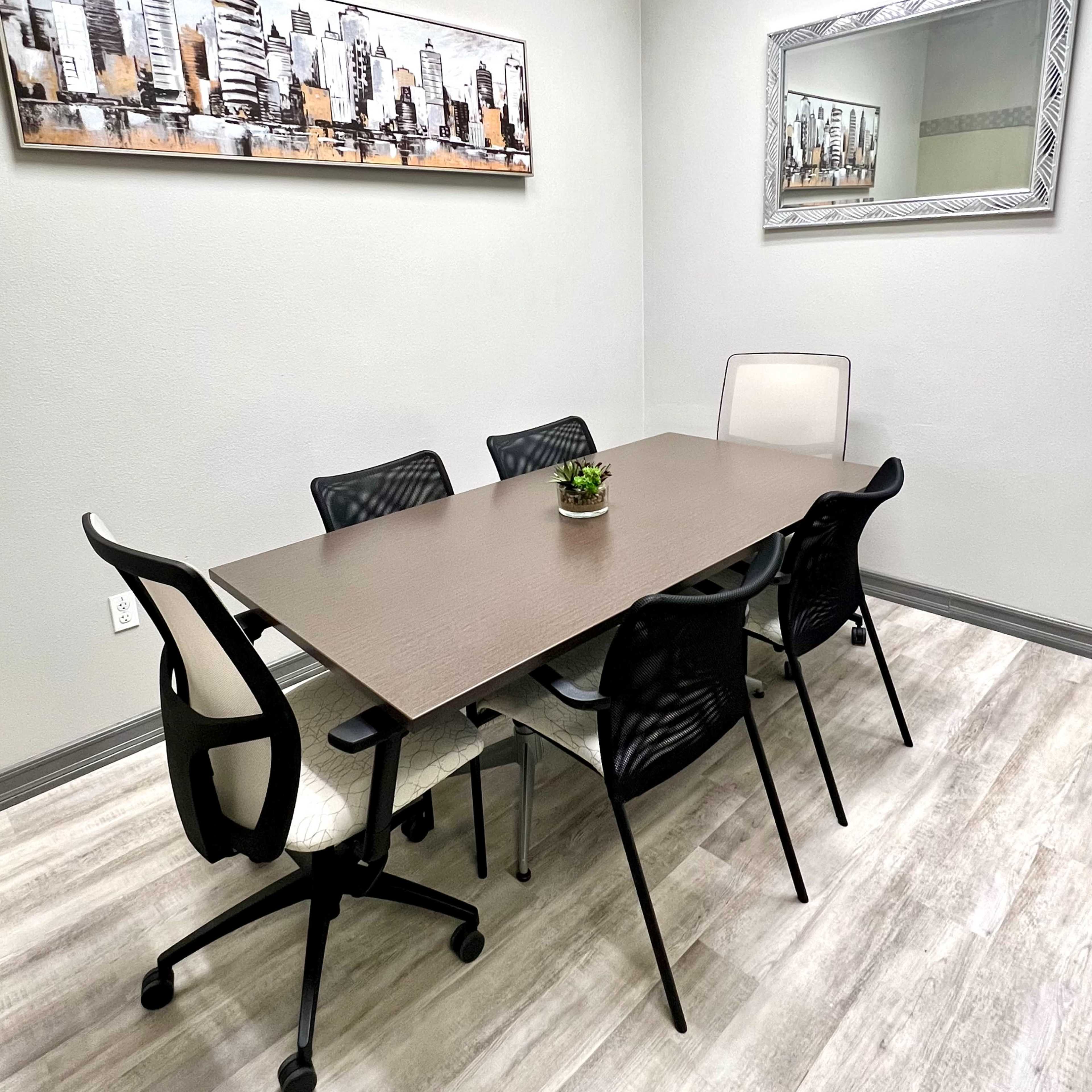 A small conference room with a rectangular table surrounded by six chairs, a wall-mounted cityscape art piece, and a mirror.