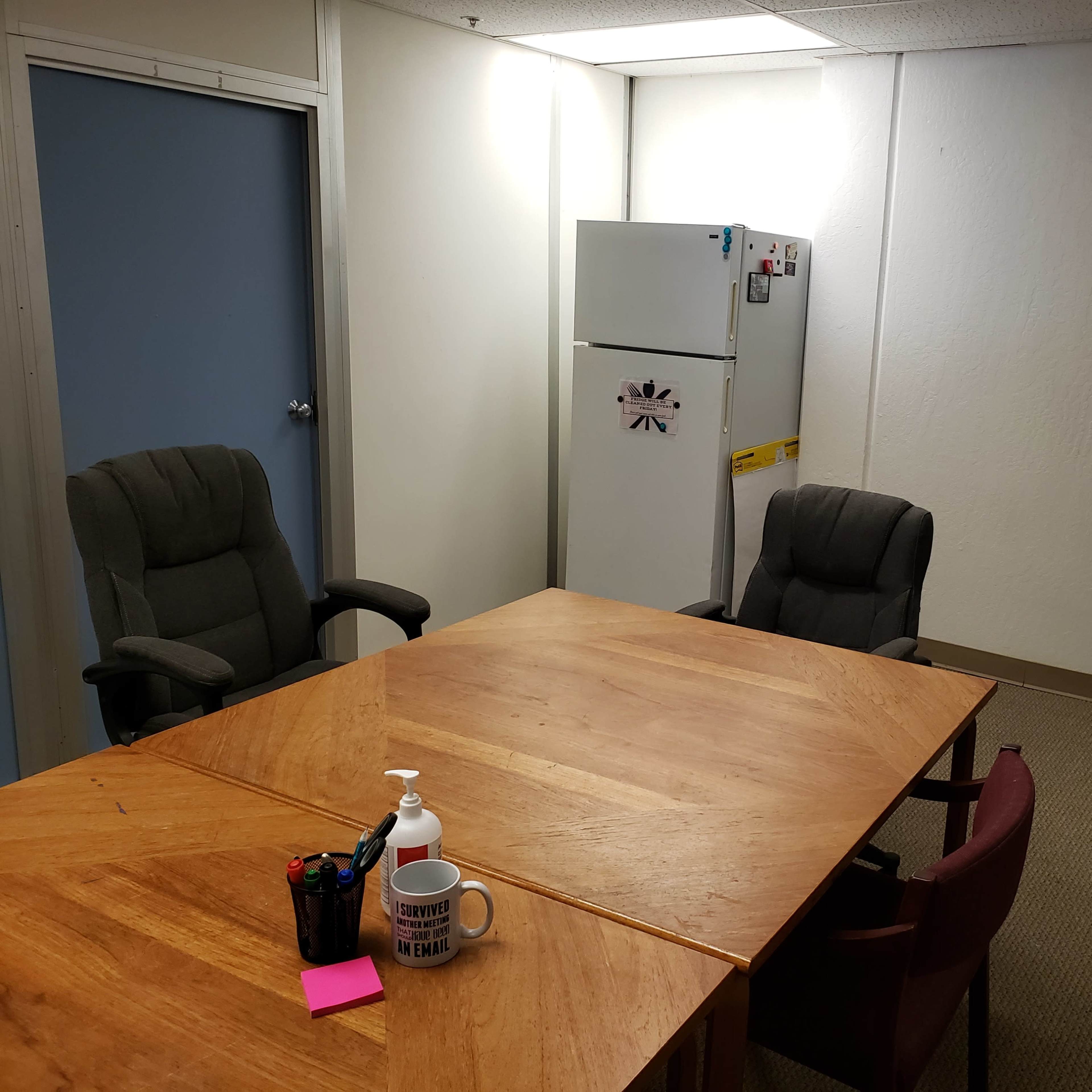 A small empty meeting room features a wooden table, two office chairs, a refrigerator, and a shelf with supplies.