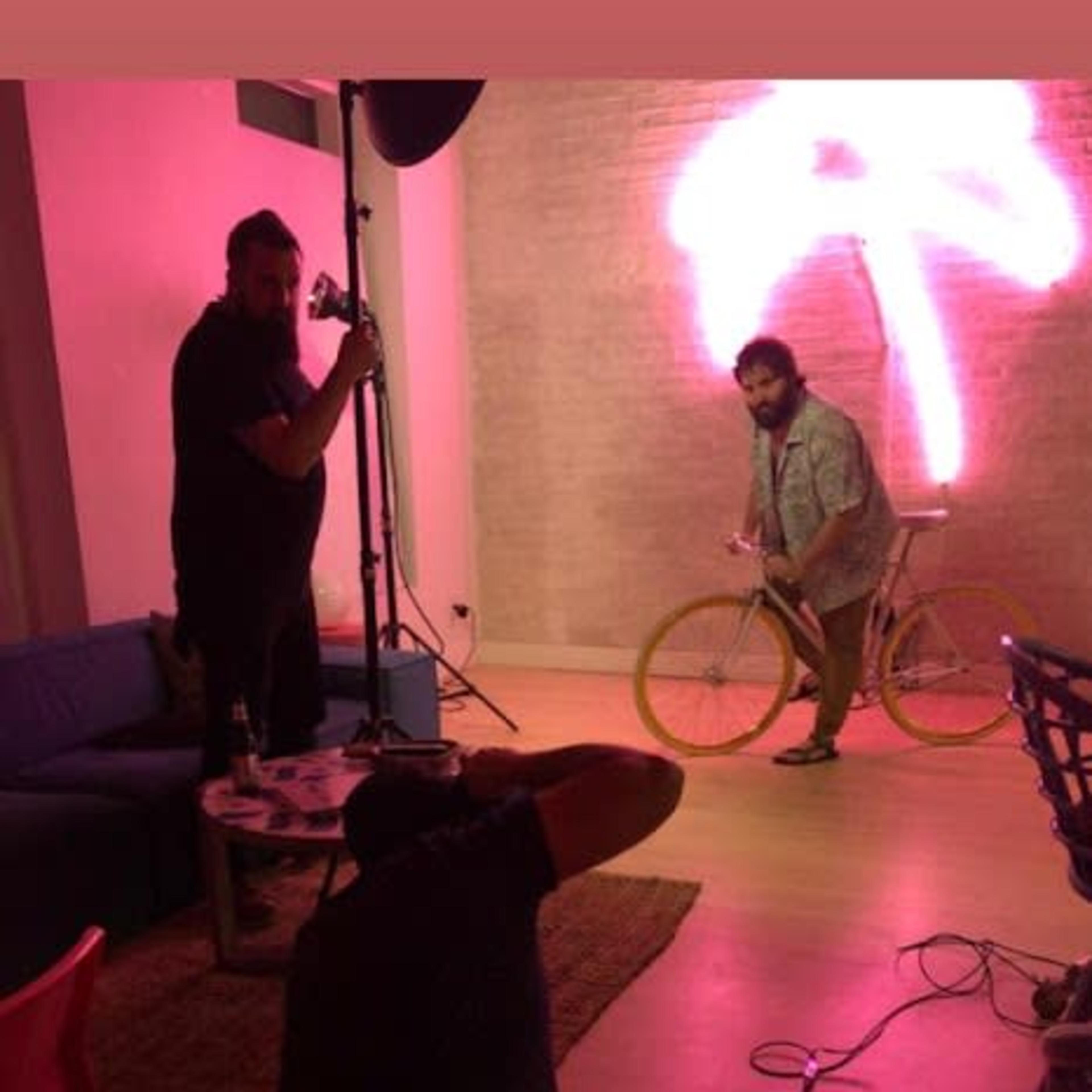 A man is posing on a bicycle in a room with pink neon lighting while another man is taking photographs.