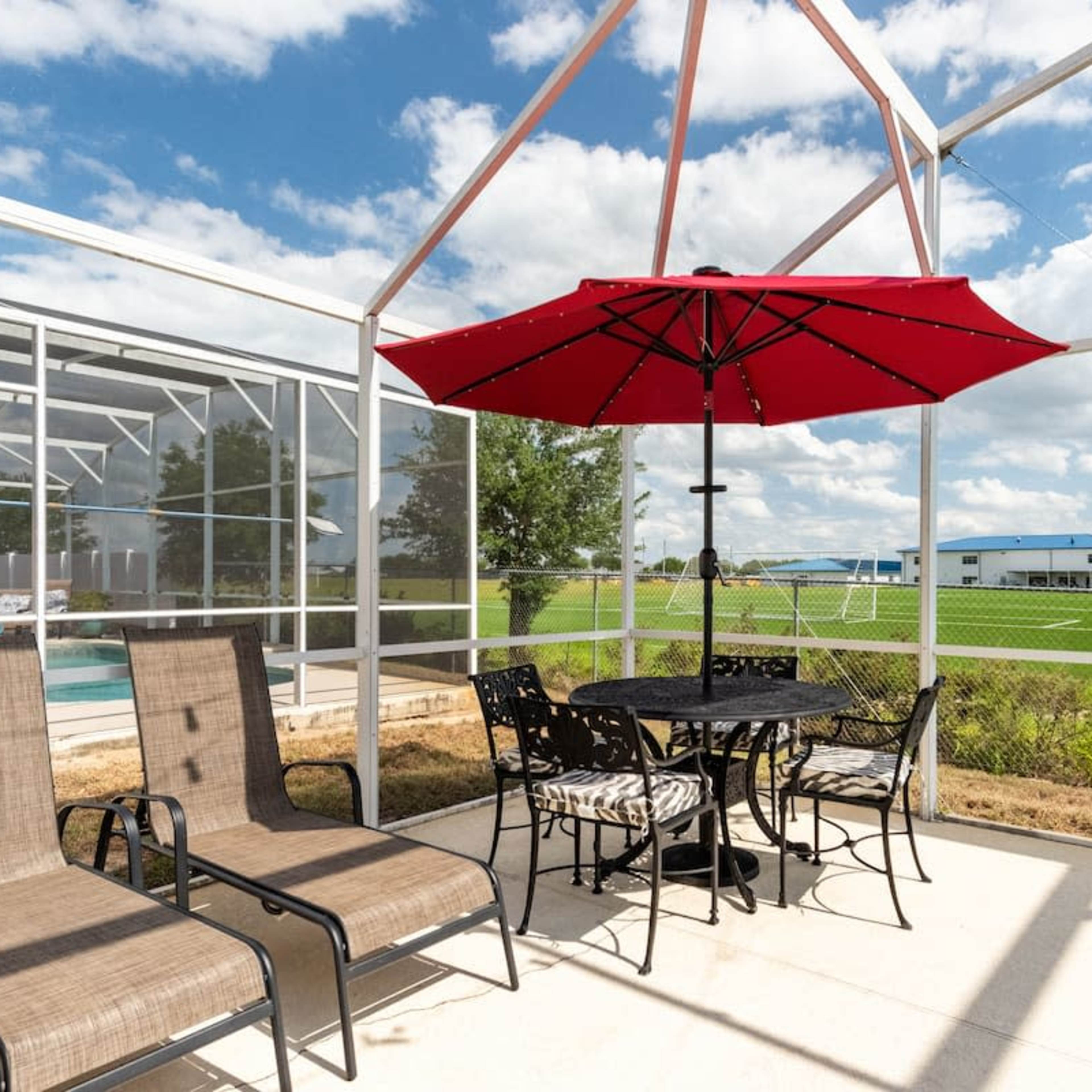 A patio area features a round table with chairs under a red umbrella, surrounded by lounge chairs, with a view of a grassy field and a building in the background.