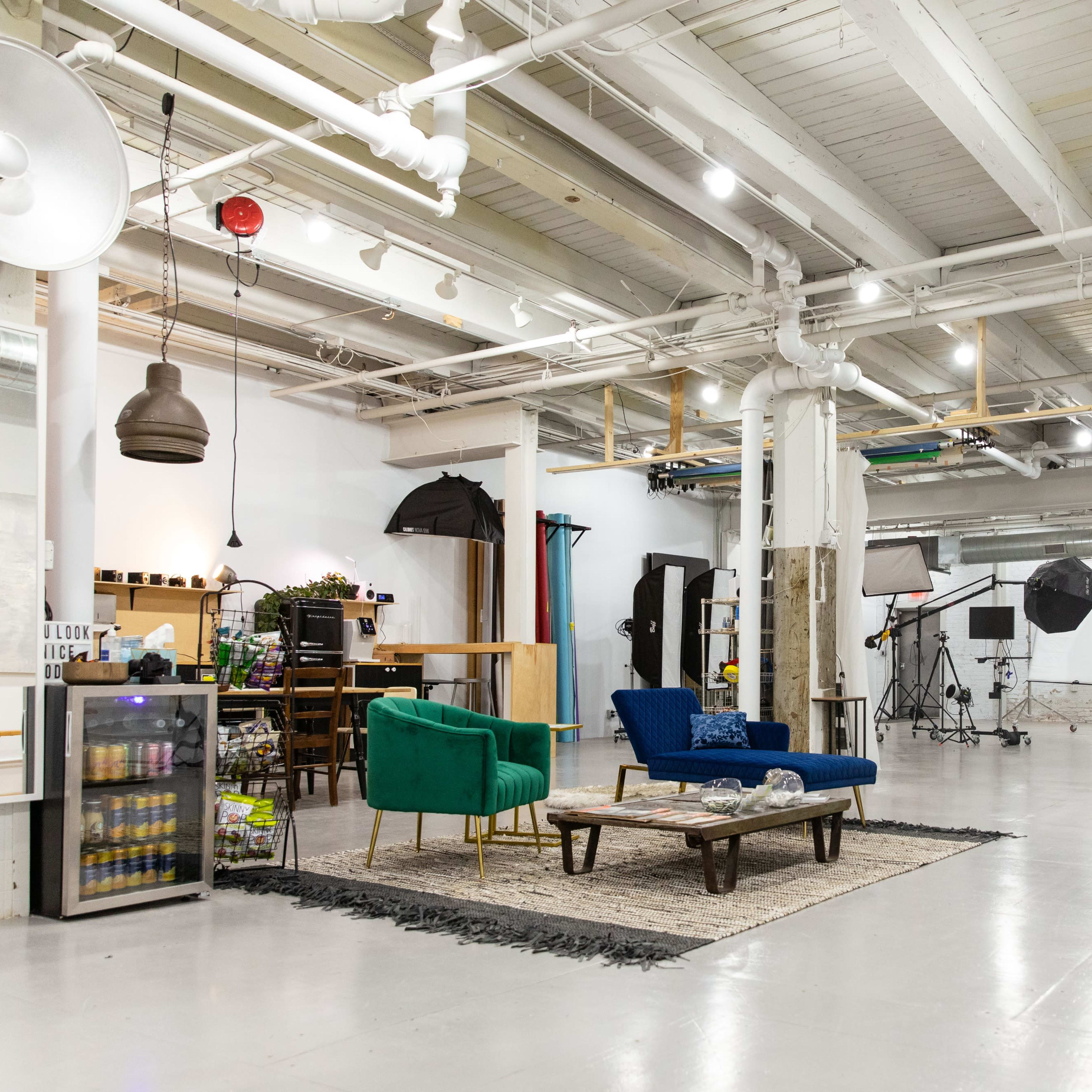 A spacious studio designed for photography, featuring seating areas with blue and green sofas, various lighting equipment, and sets in the background.