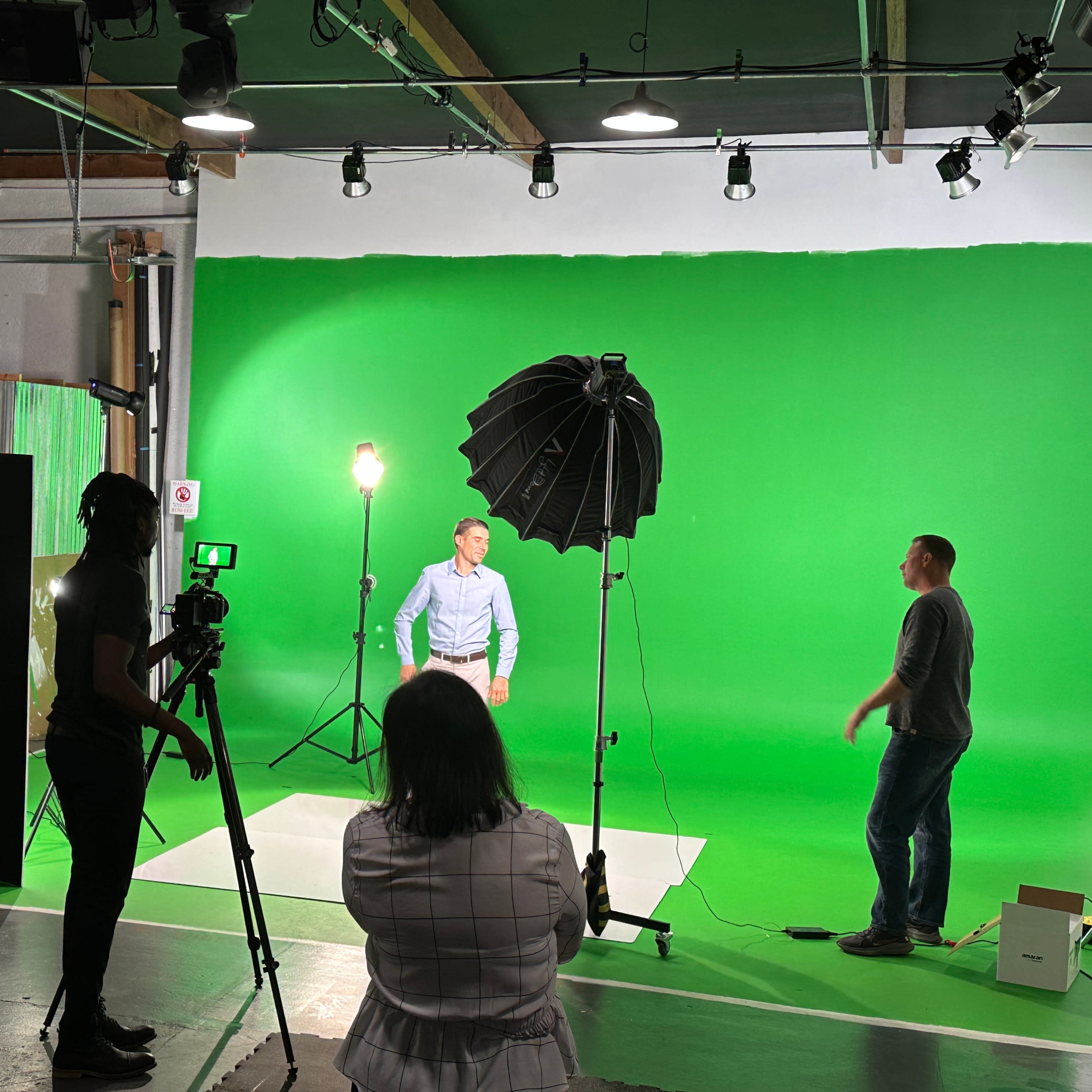 A film crew sets up a green screen studio with multiple lights and a camera while two individuals prepare for a shoot.