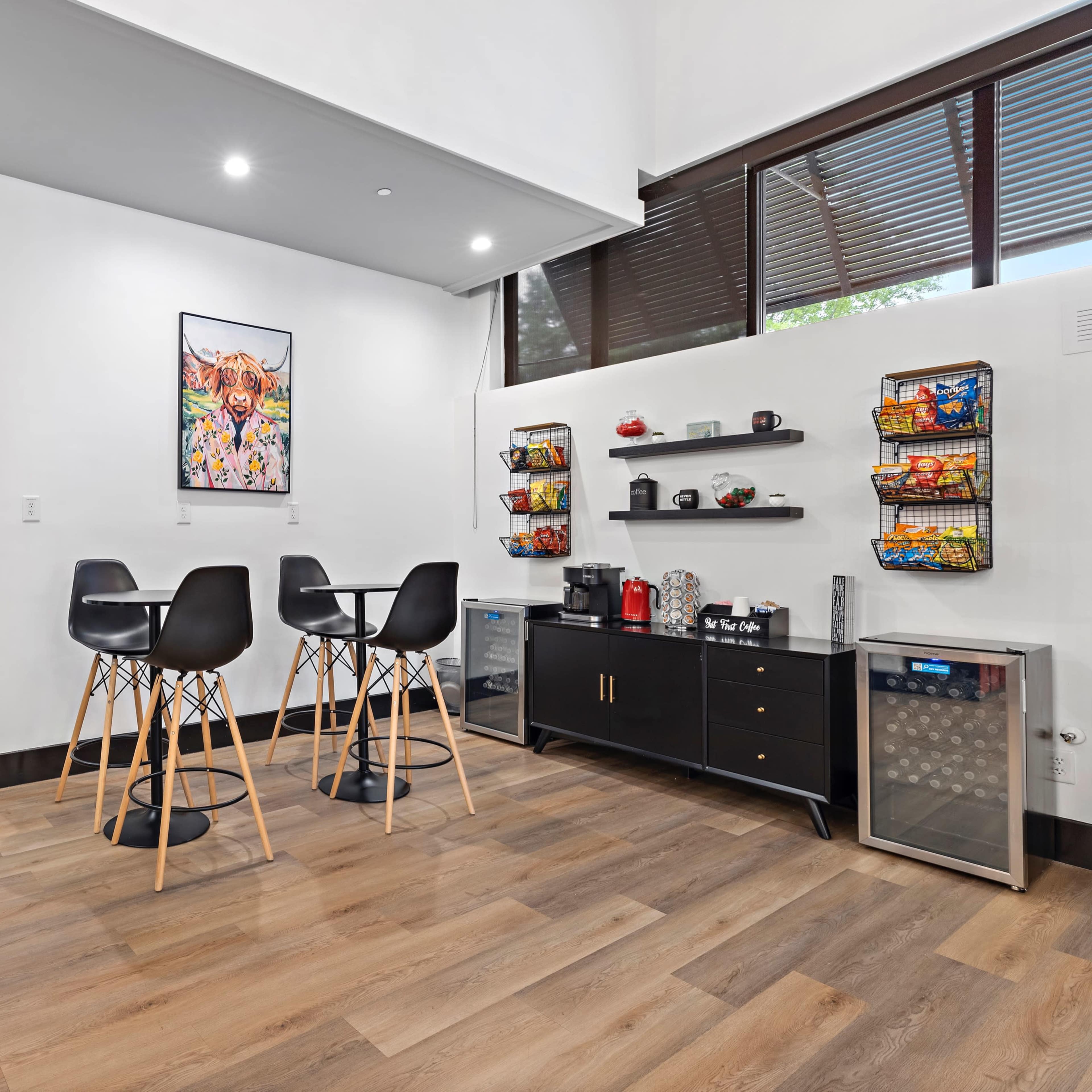 A modern common area with a small dining table and chairs, a kitchenette with snacks, and a corridor leading to multiple closed doors.