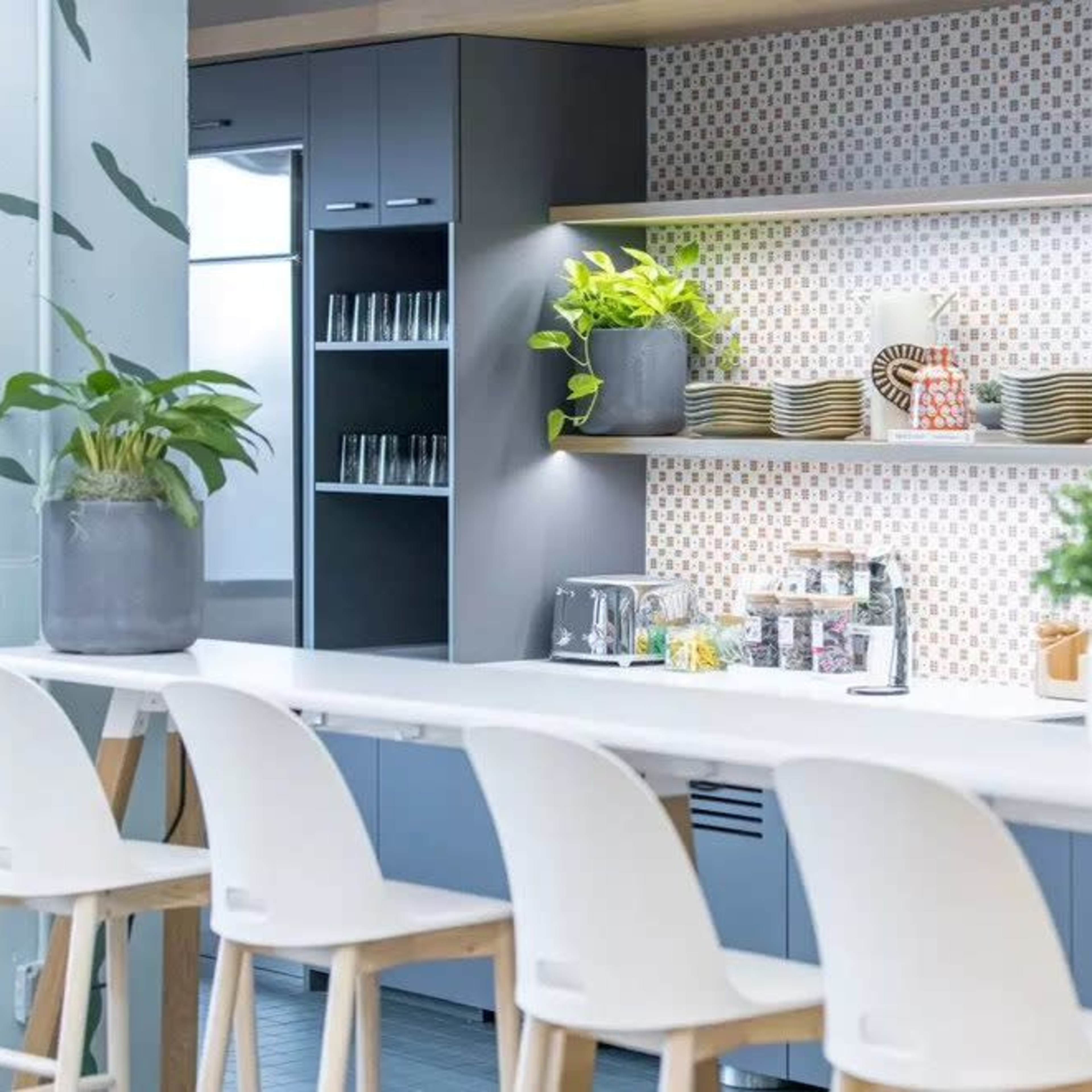A modern kitchen space features a long countertop with bar stools, a row of plants, beverage dispensers, and neatly arranged dishes against a patterned backsplash.