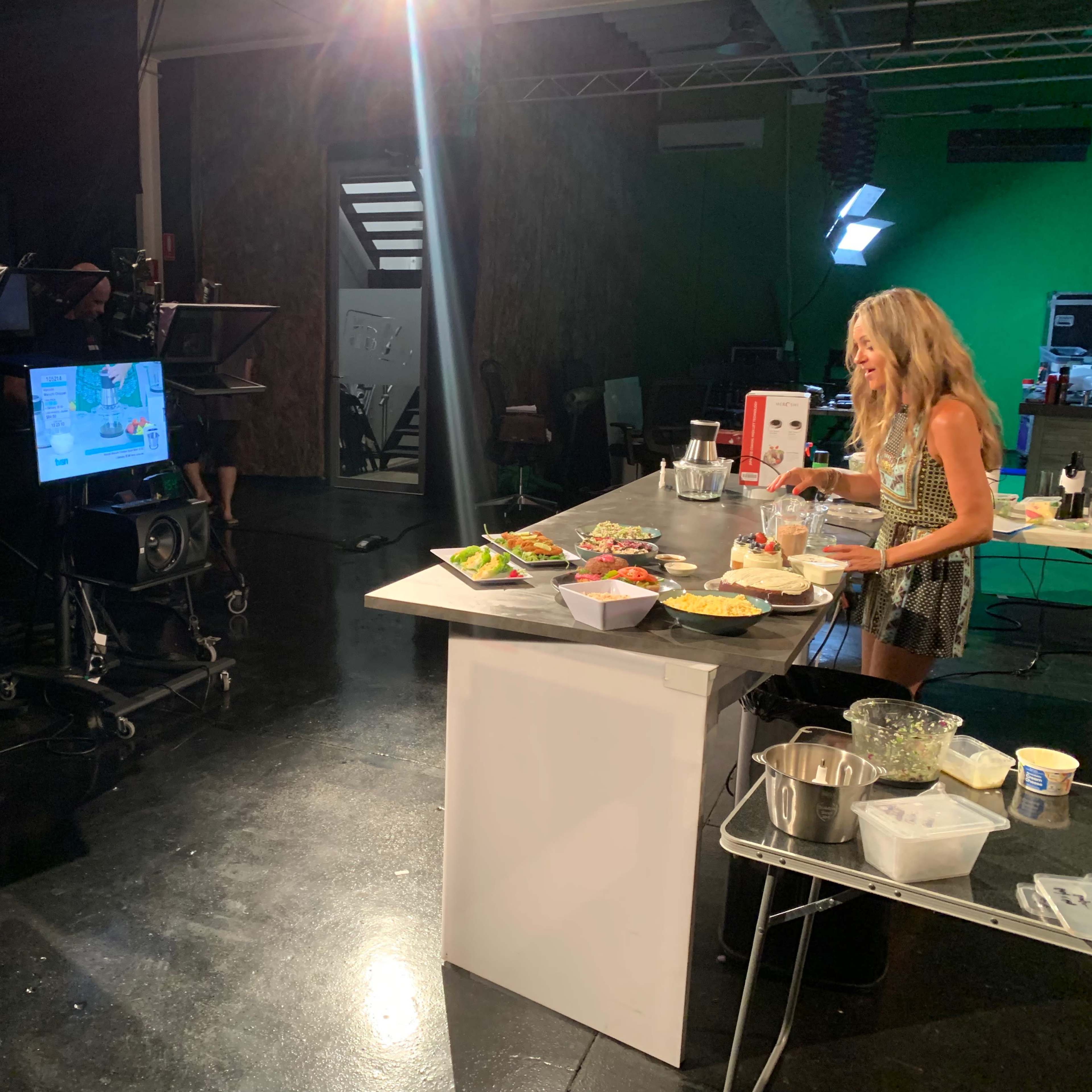 A woman prepares food at a studio kitchen setup, with cameras and screens positioned around her.