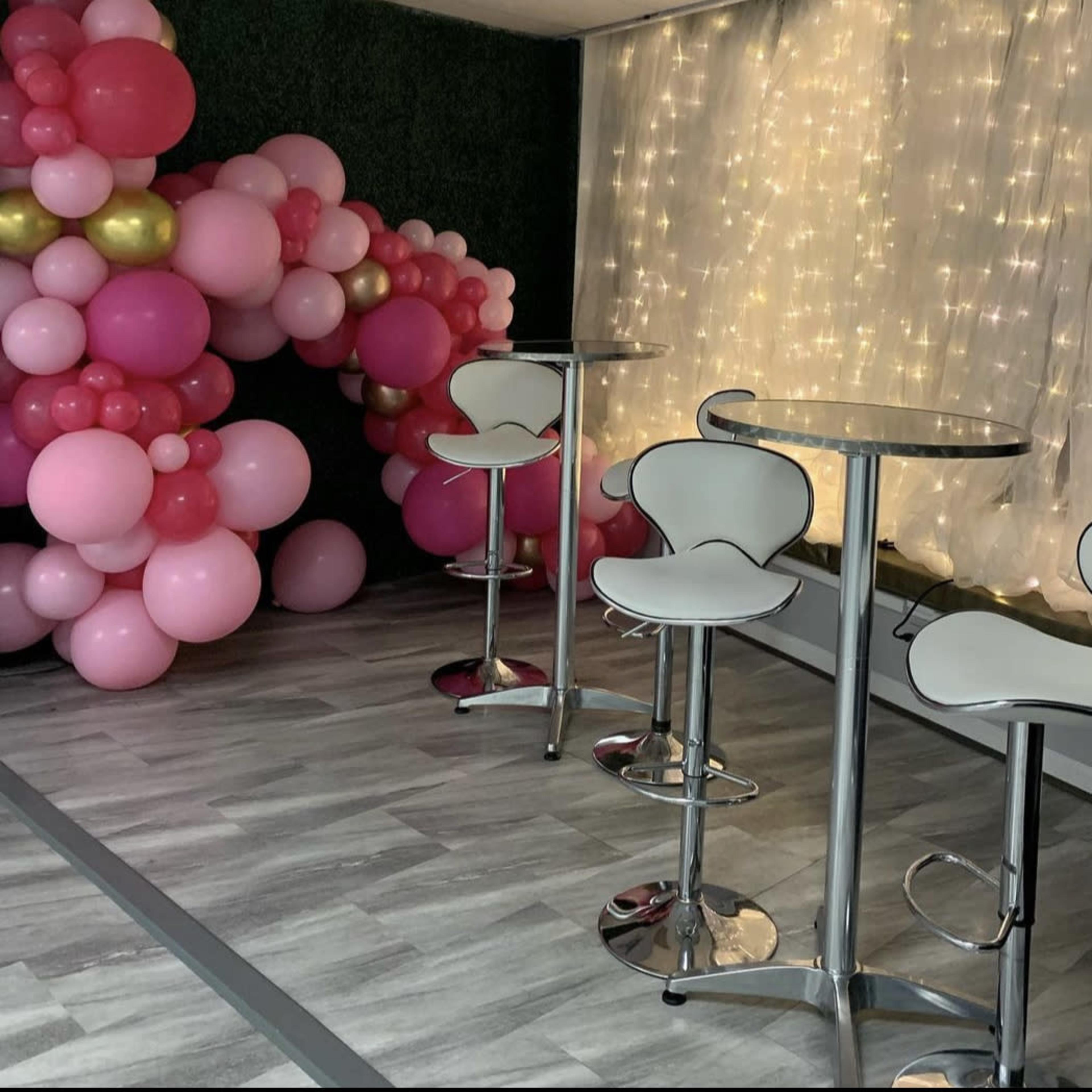 The scene features a small event space with three white bar stools and tables, alongside a decorative arrangement of pink and gold balloons against a backdrop of twinkling lights.