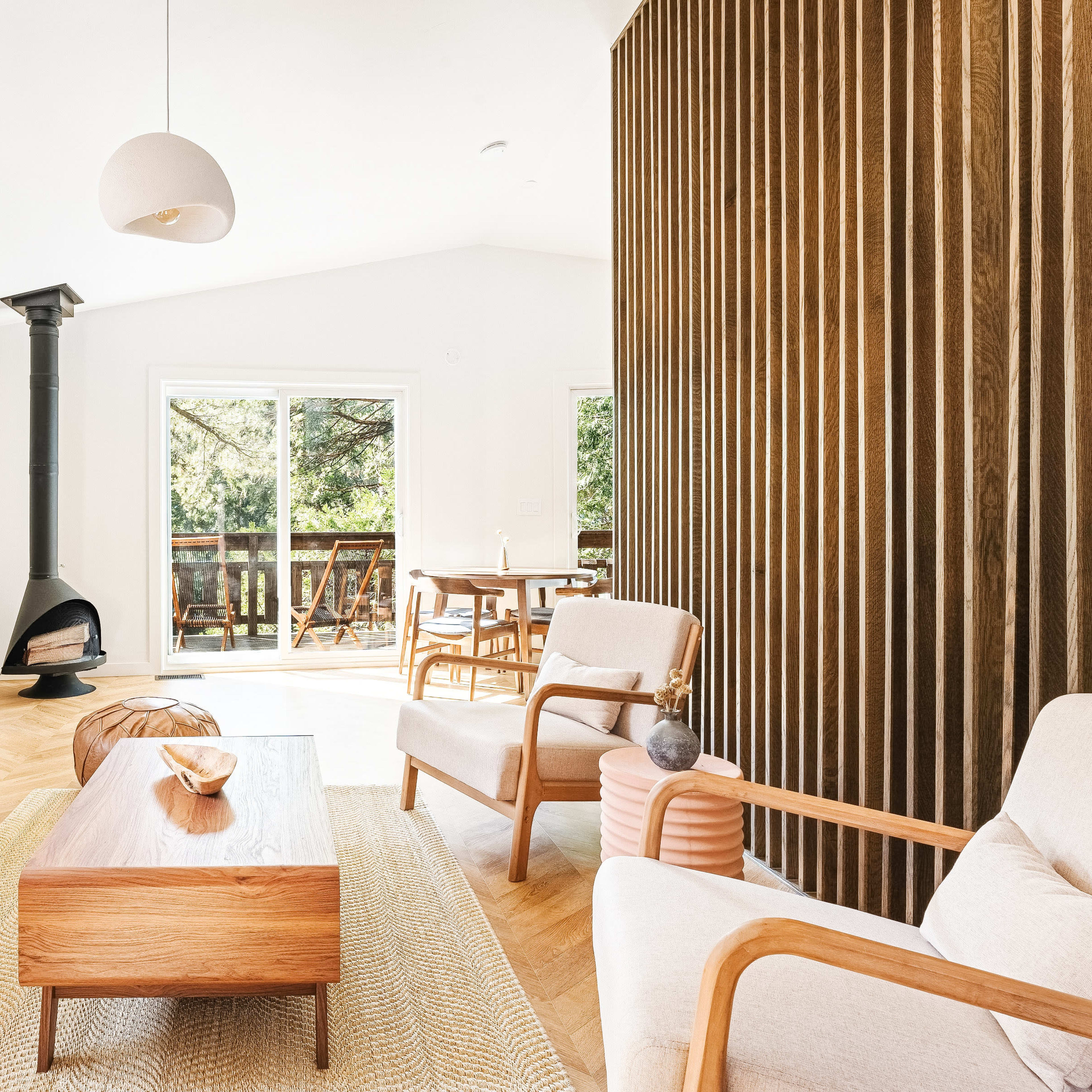 A modern living room features a brown leather sofa, a wooden coffee table, and a wood-burning stove, with large windows that overlook a deck surrounded by trees.