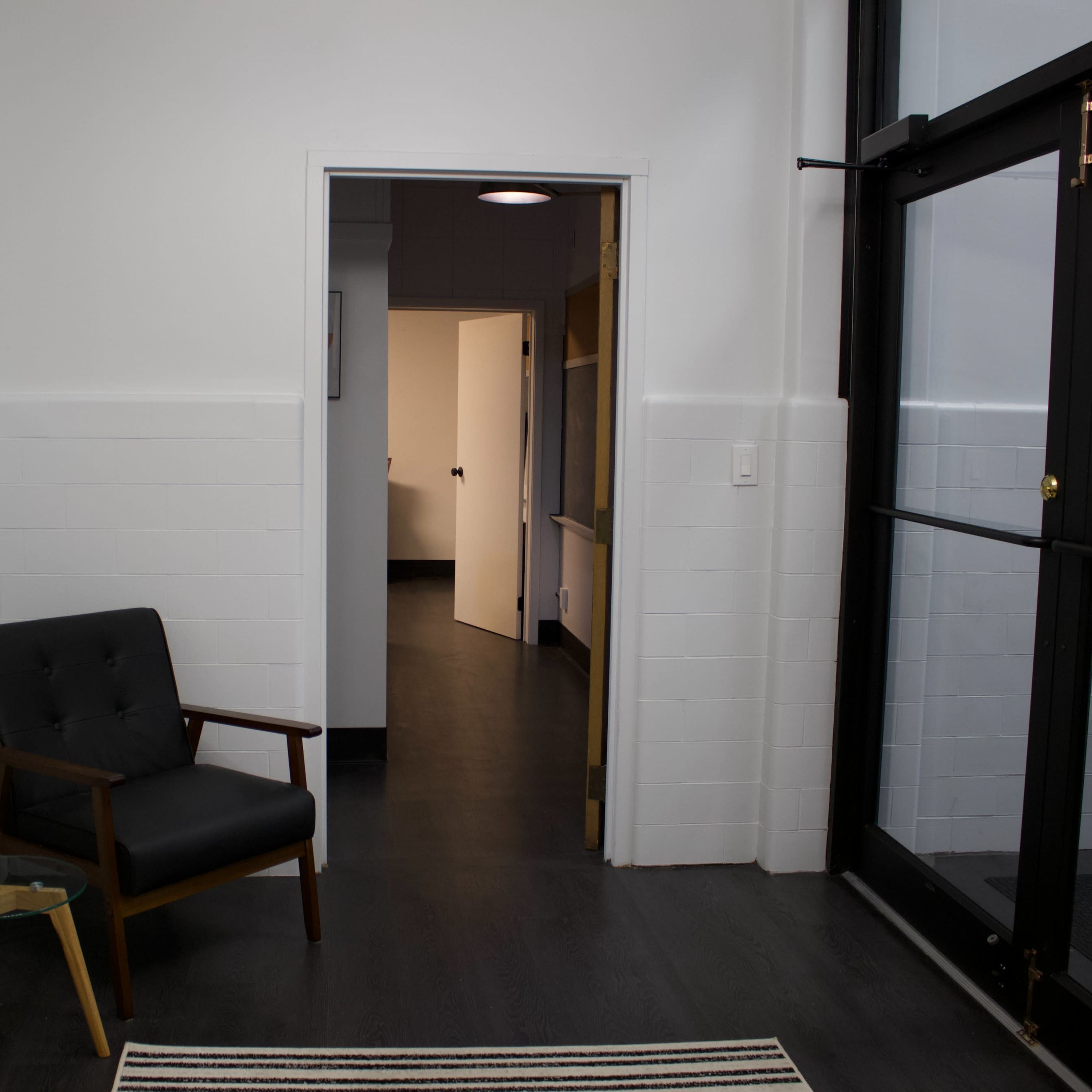 A minimalist entryway featuring a black chair next to a small table, with a door leading to a hallway in the background.