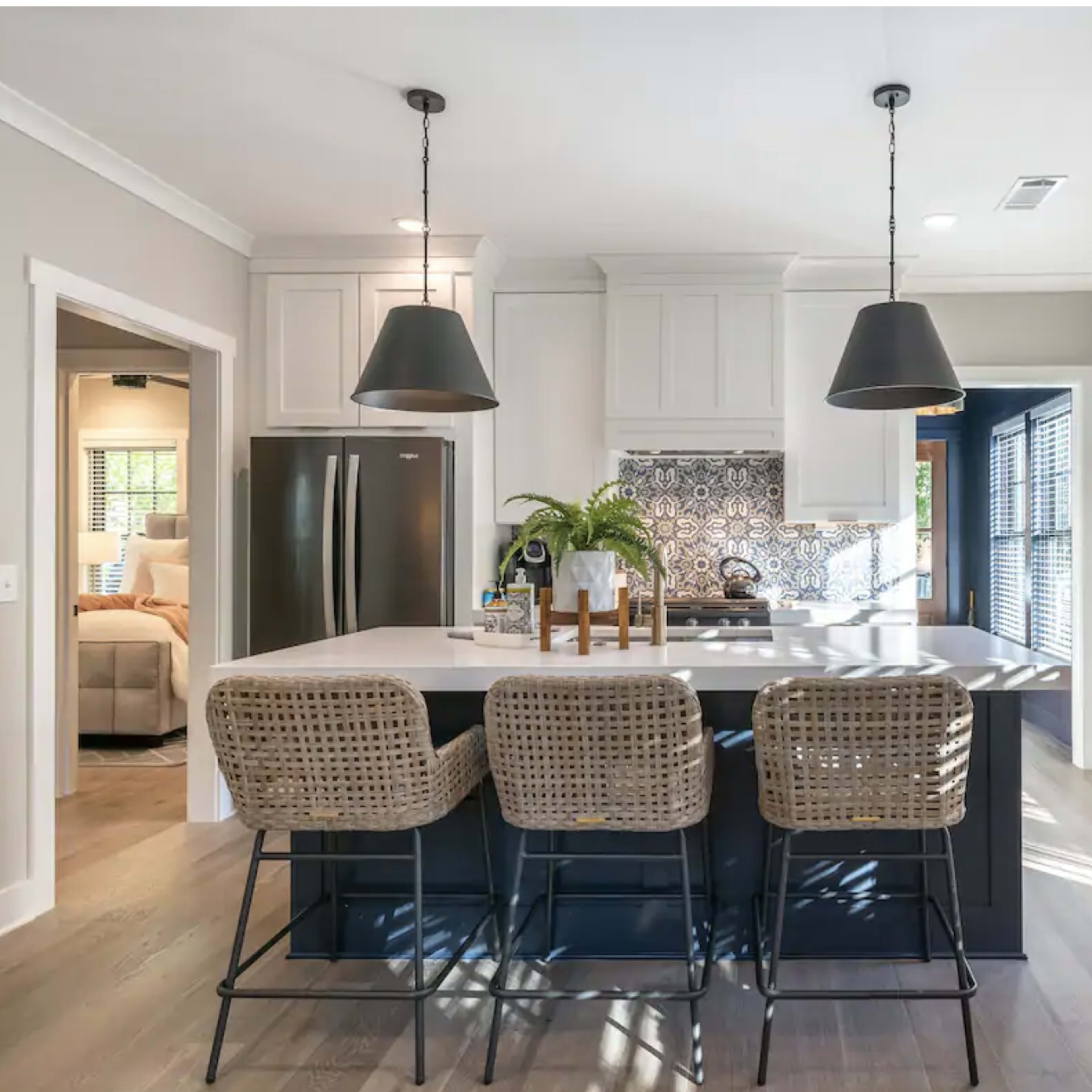 A modern kitchen with a central island featuring three wicker bar stools, pendant lights above, and a patterned backsplash, adjacent to a doorway leading to a living area.