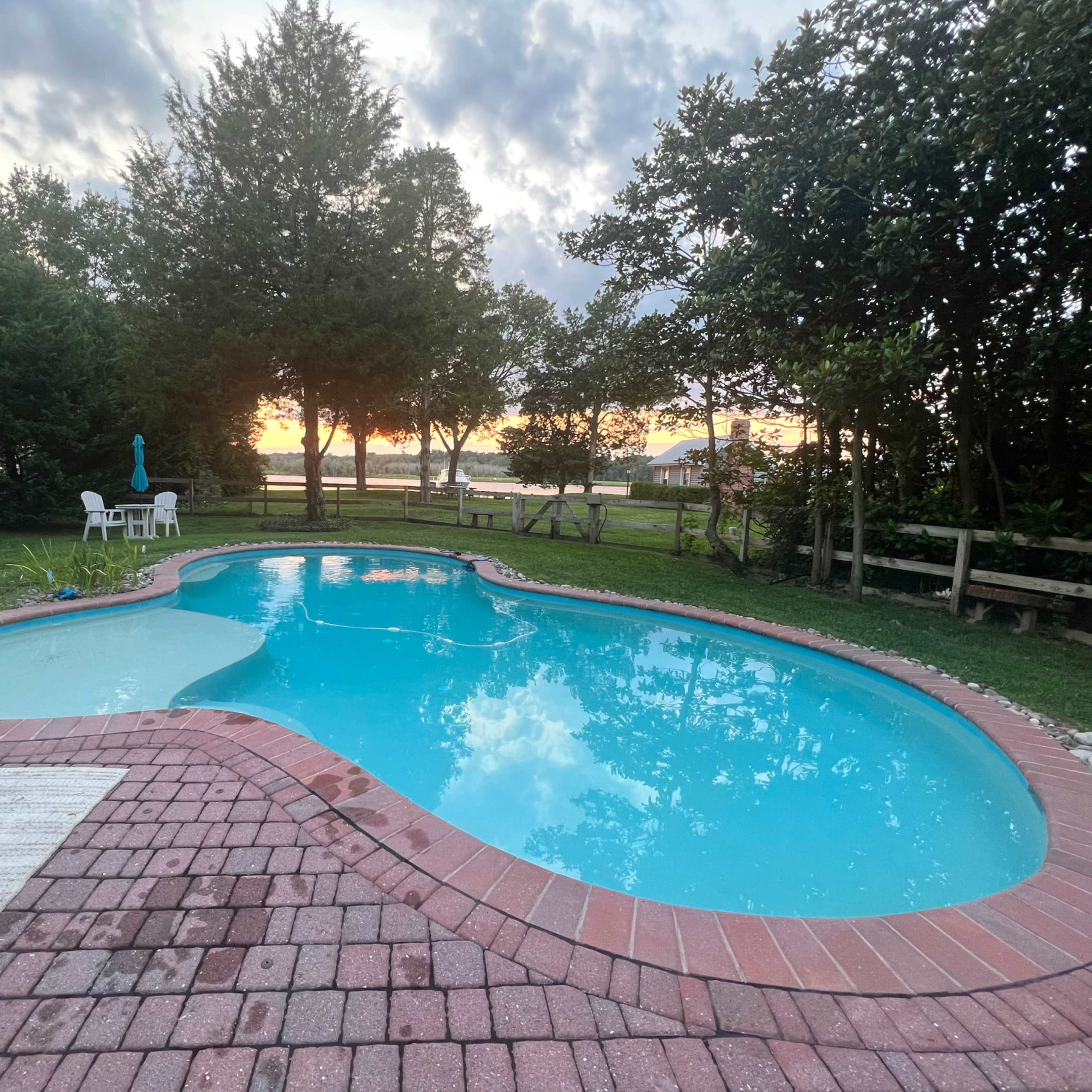 A swimming pool with a brick patio surrounded by greenery, set against a sunset backdrop.