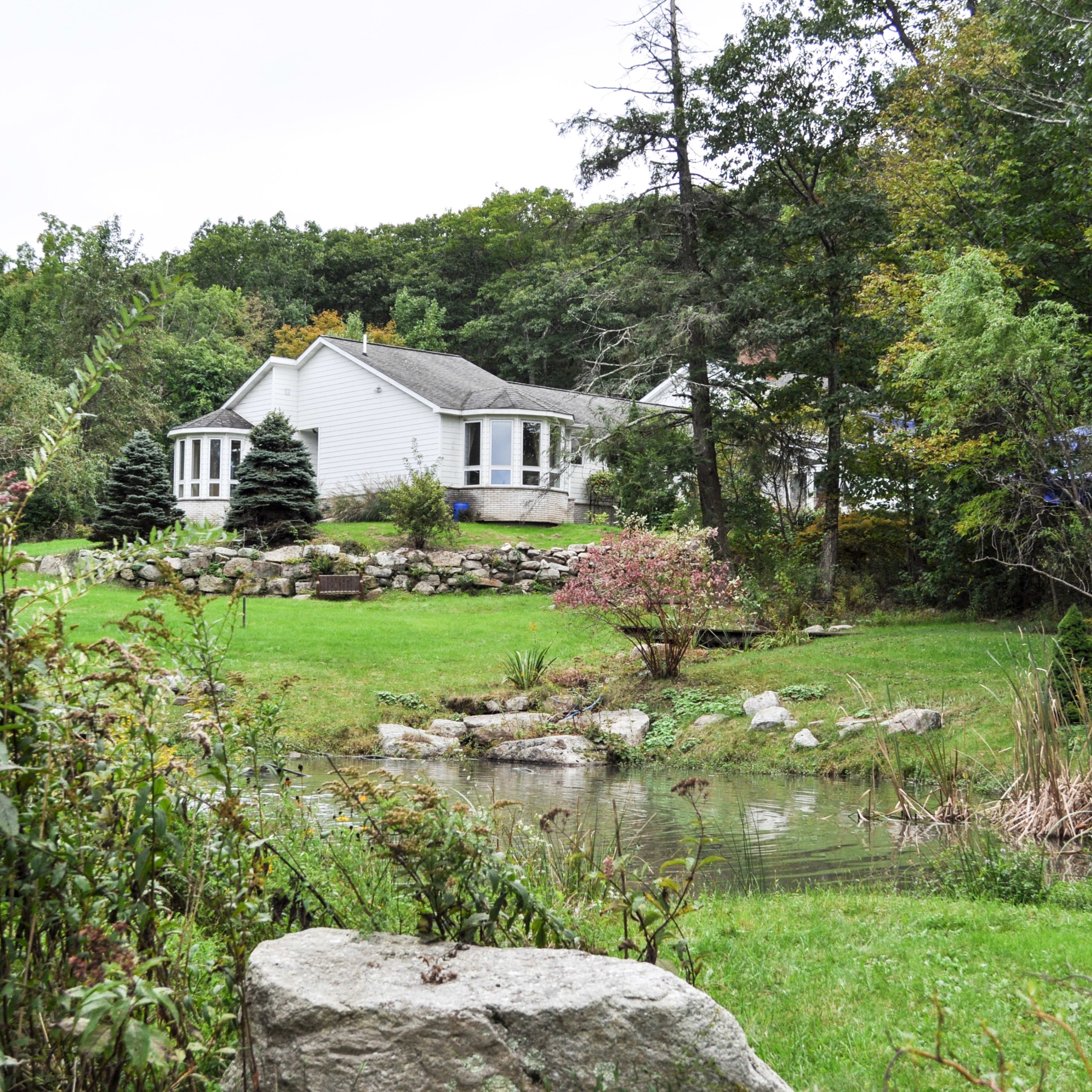 A white house is situated near a pond, surrounded by lush greenery and landscaping in a wooded area.