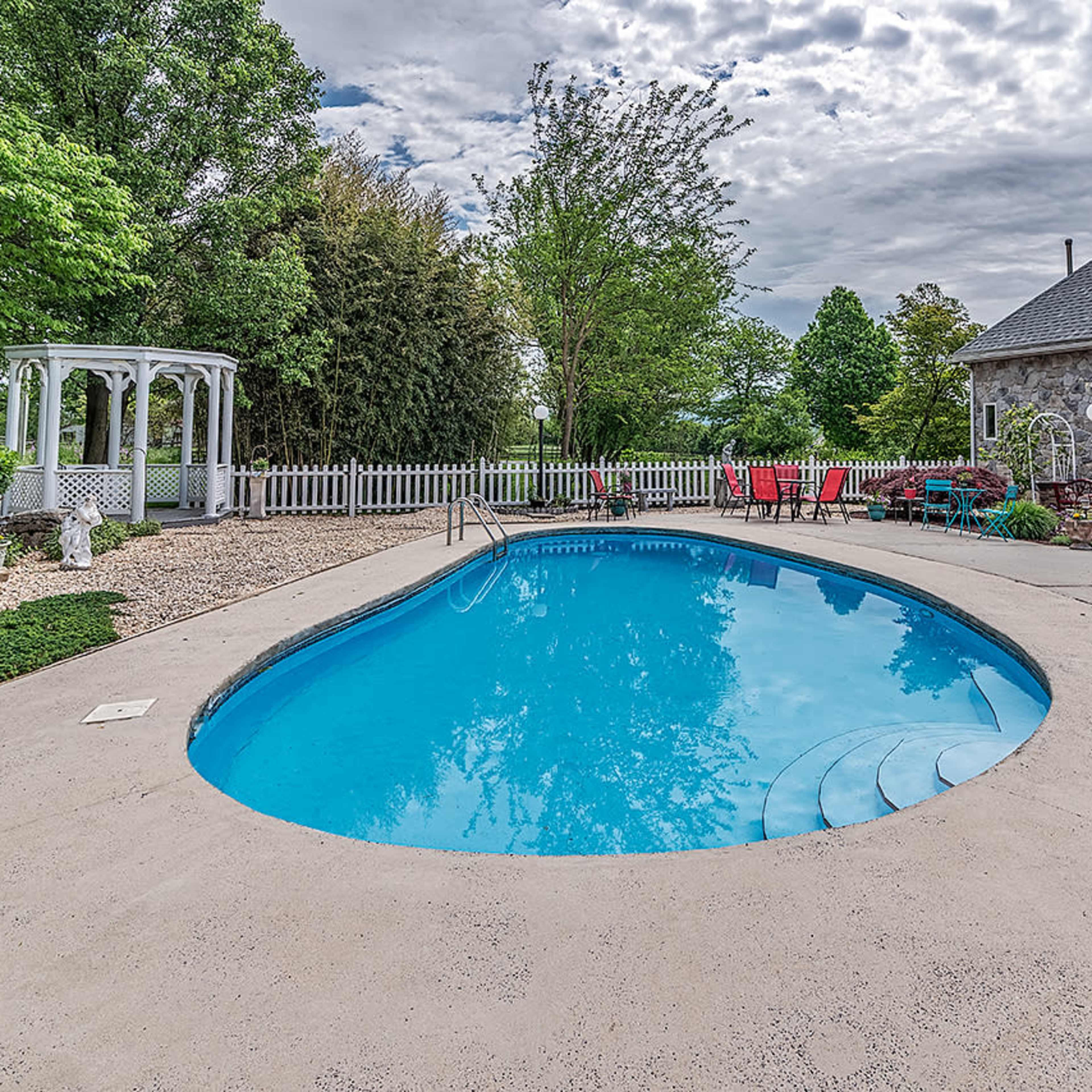 A swimming pool surrounded by a patio, garden furniture, and a gazebo next to a stone house.