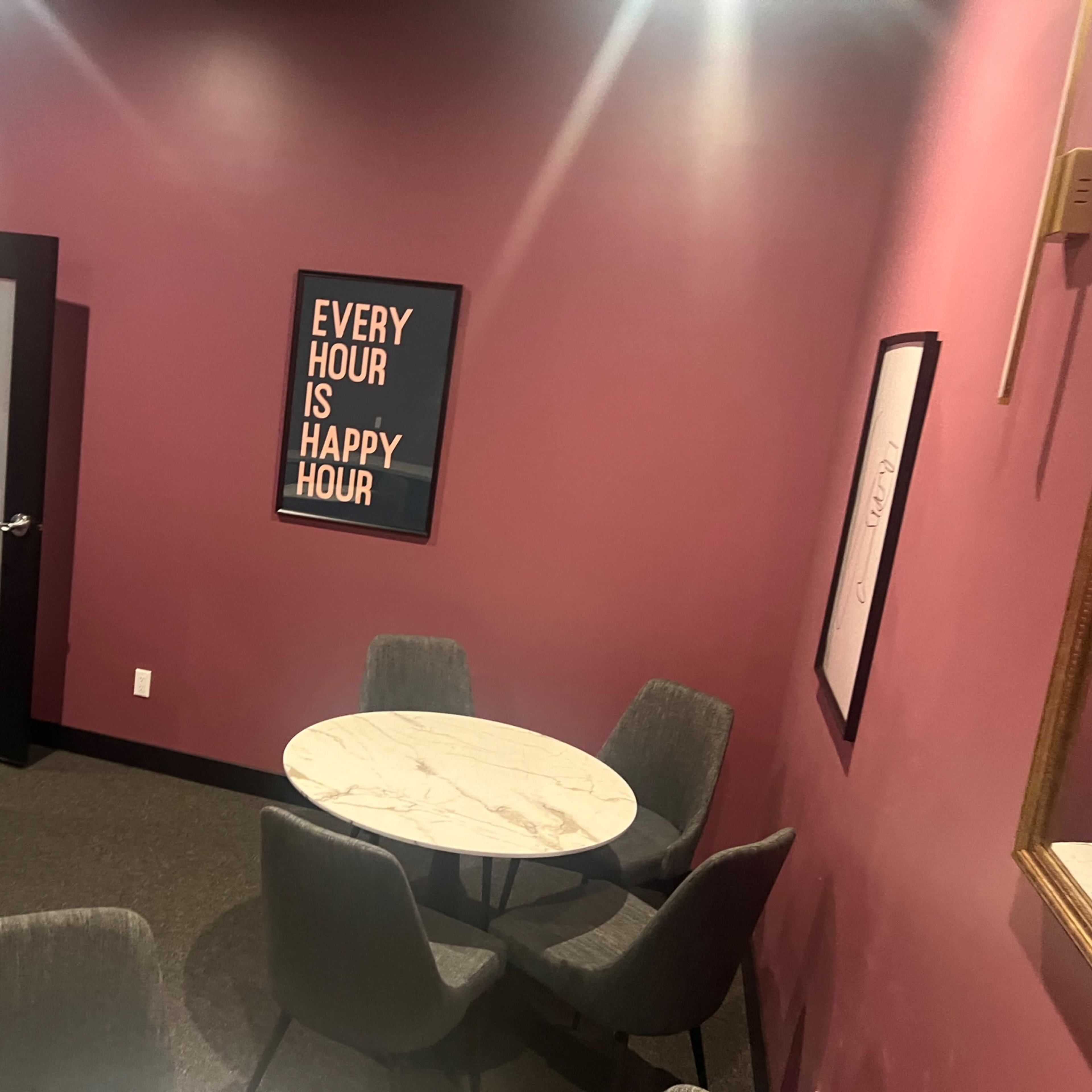 A small room features several round marble tables surrounded by gray chairs, with a pink wall and a television mounted above a countertop.
