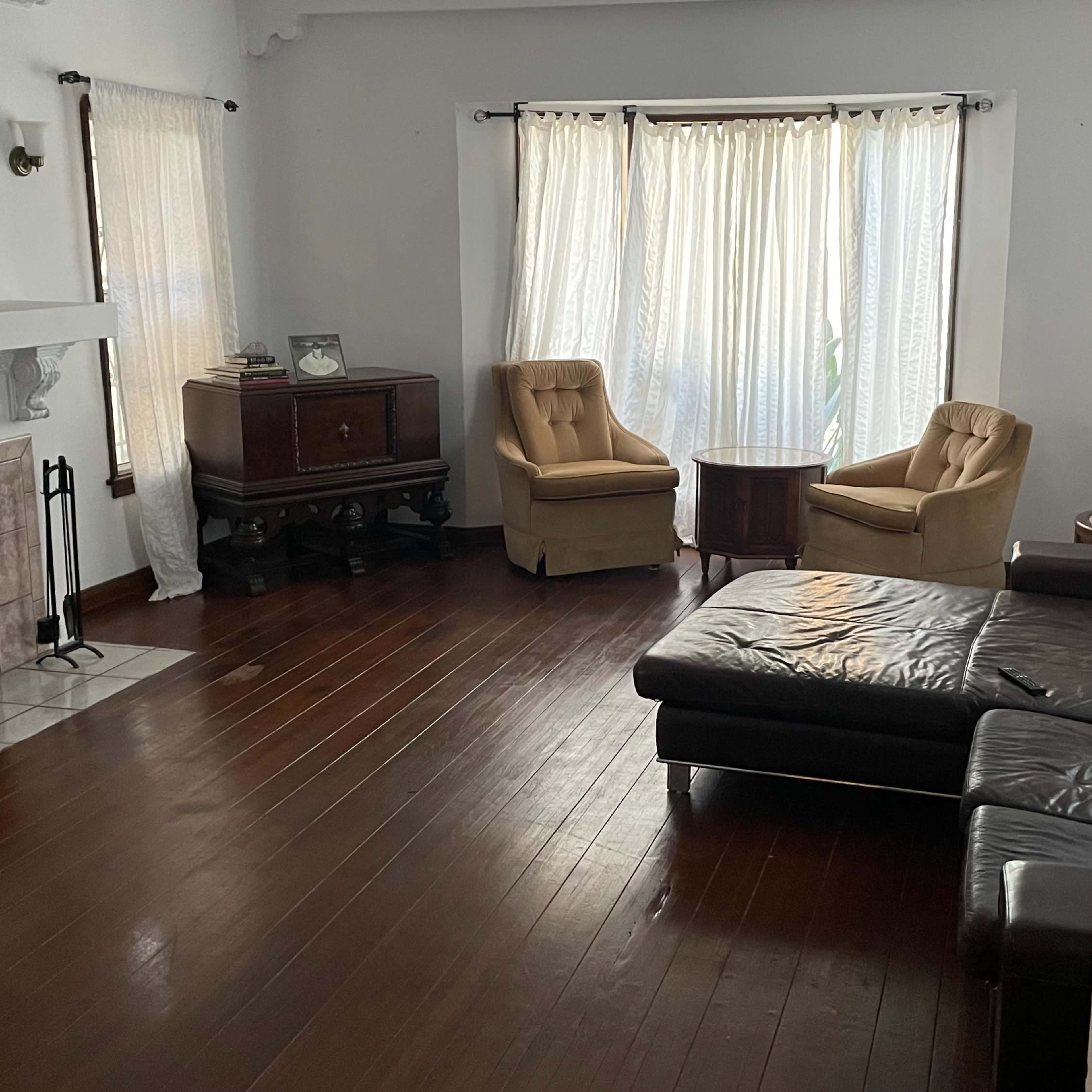 A living room features a television on the wall, two armchairs, a small table, and a leather sectional sofa, with natural light streaming through sheer curtains.