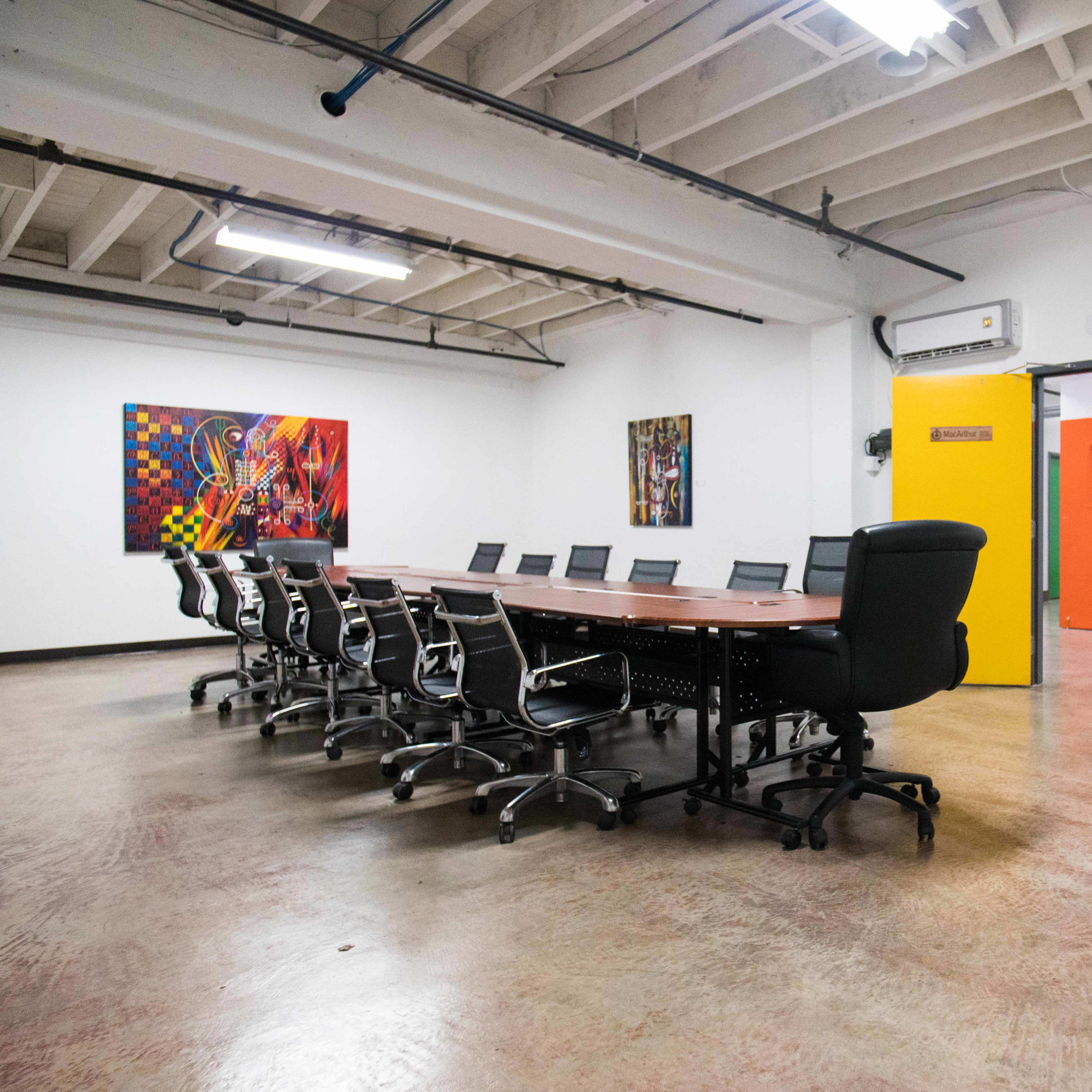 A large conference room features a long table surrounded by ergonomic chairs, with colorful artwork on the walls and an orange door leading to another area.