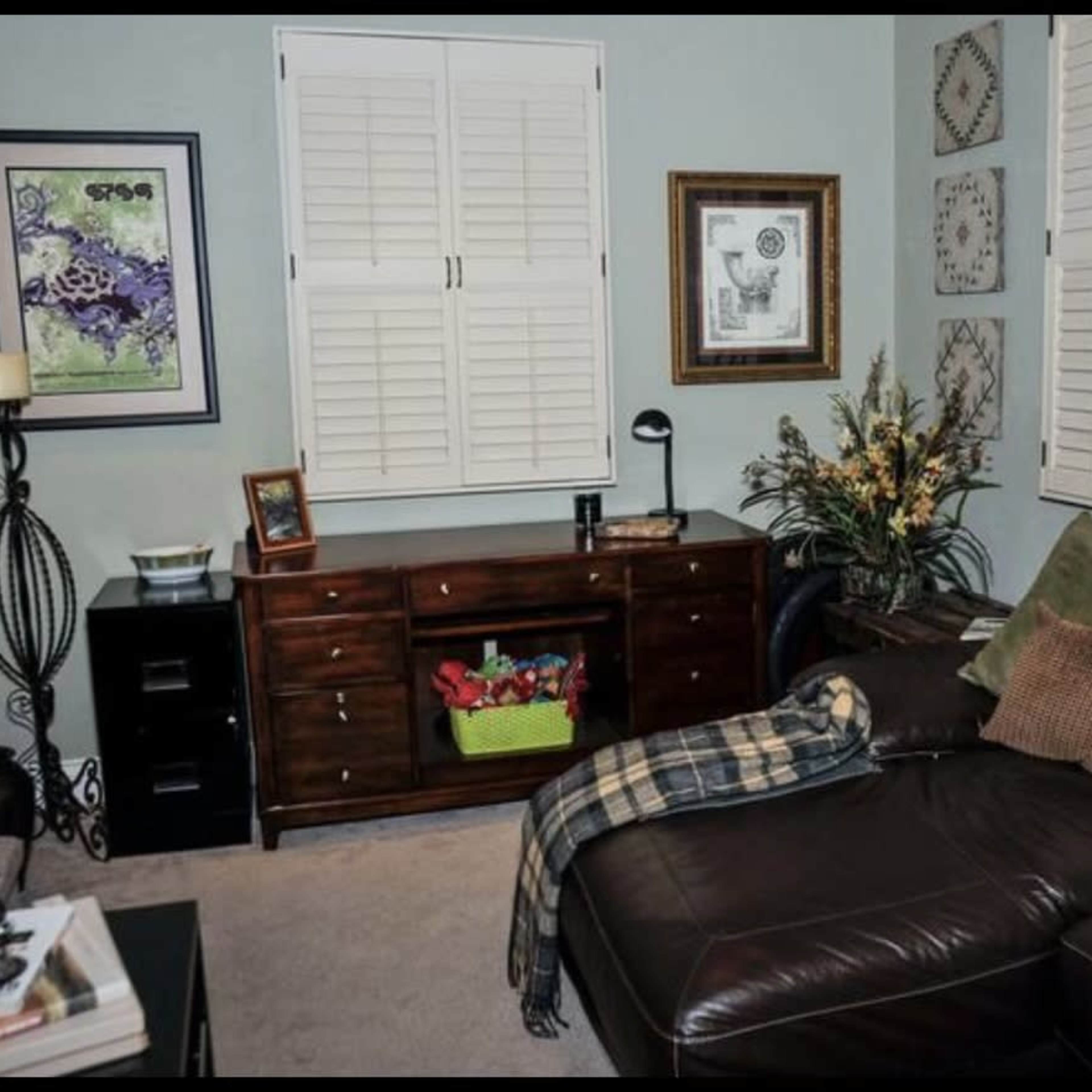 The room features a brown leather sectional sofa, a wooden cabinet with decorative items, and light blue walls adorned with framed art and plants.